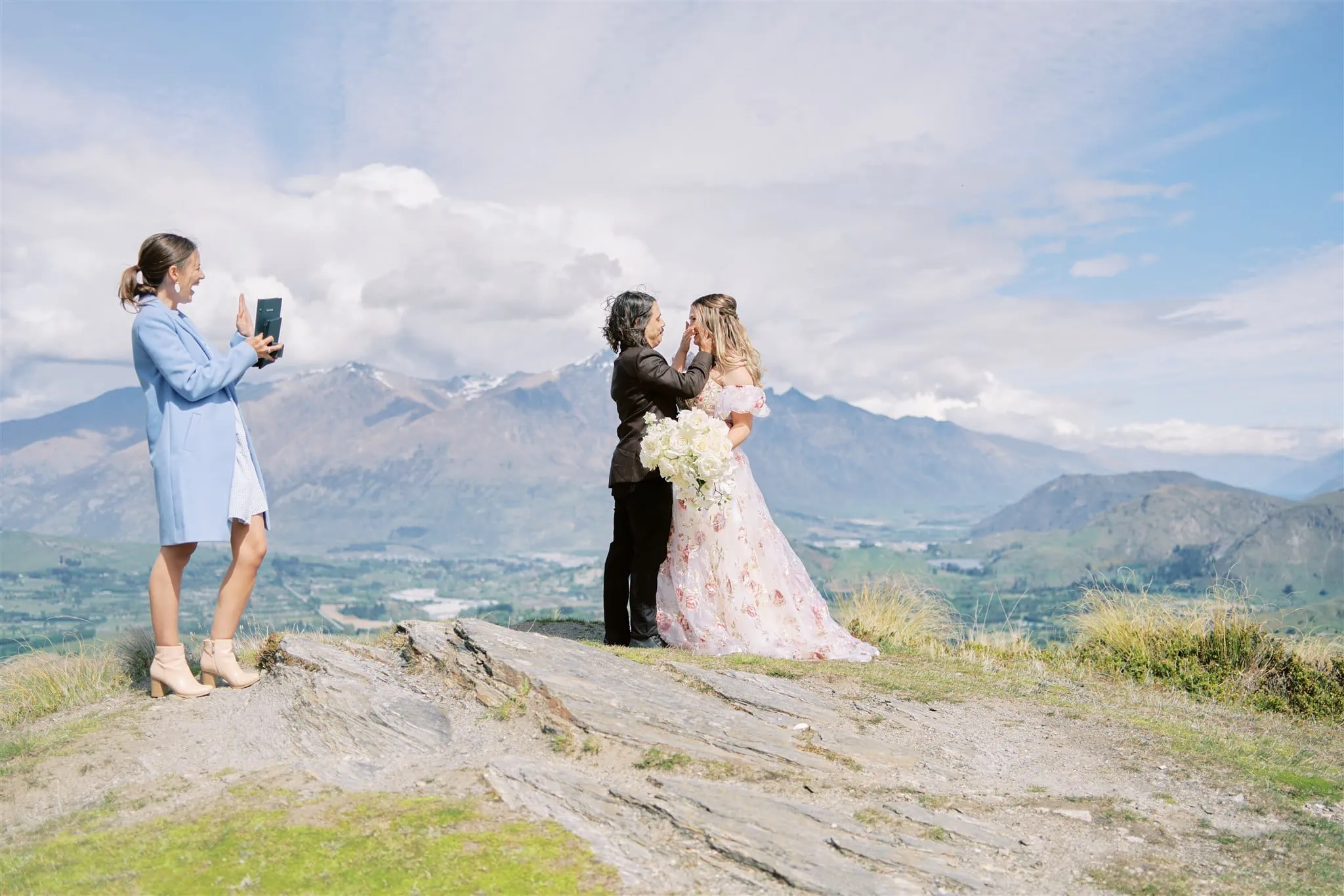 Queenstown Tekapo New Zealand Heli Wedding Elopement Pre-Wedding Shoot Photographer クイーンズタウン　テカポ　ニュージーランド　エロープメント 前撮り　フォトウェディング　結婚式 | A wedding couple shares a kiss on Coronet Peak, overlooking a scenic view while a young girl captures the moment on a smartphone.