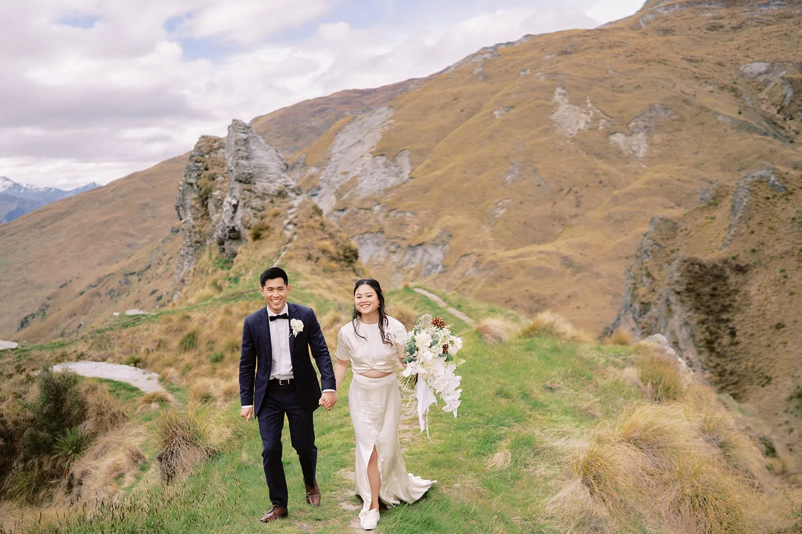 Queenstown Elopement Heli Wedding Photographer クイーンズタウン結婚式 | A couple holding hands and walking on a grassy hill for their pre-wedding photoshoot.