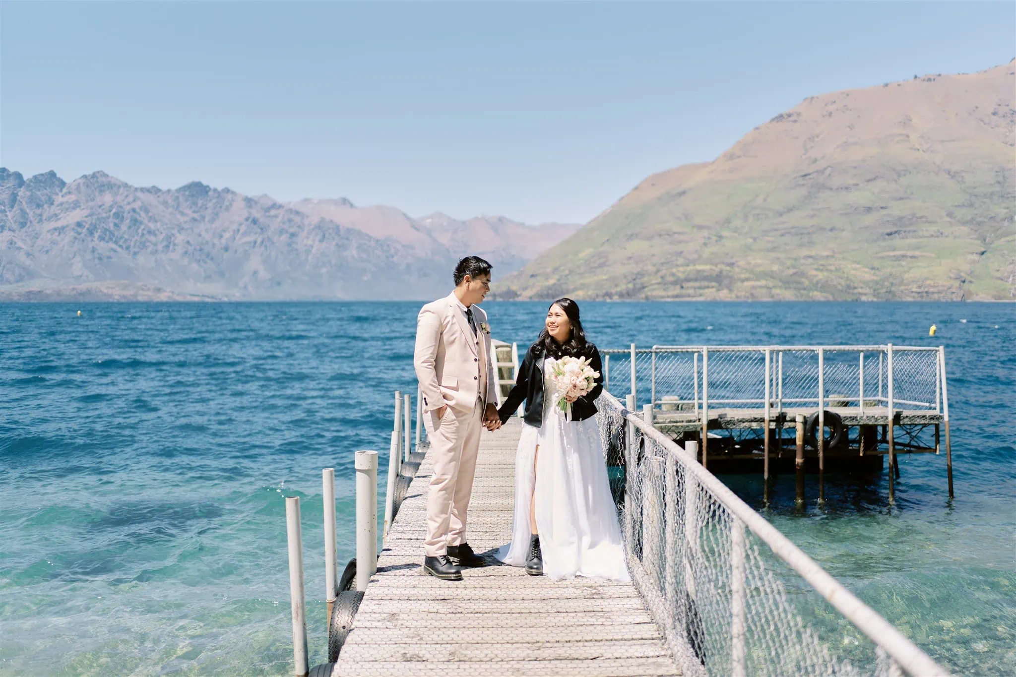 Queenstown Elopement Heli Wedding Photographer クイーンズタウン結婚式 | A Queenstown elopement with a bride and groom standing on a dock, framed by majestic mountains.