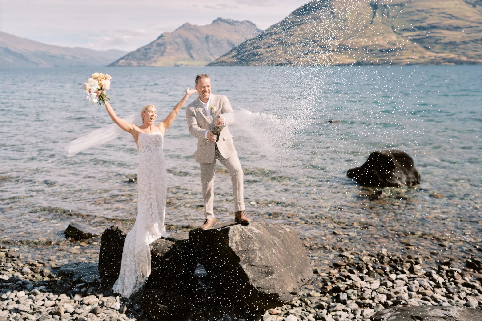 Queenstown Elopement Heli Wedding Photographer クイーンズタウン結婚式 | A Queenstown elopement with a bride and groom standing on rocks in front of a lake in New Zealand.