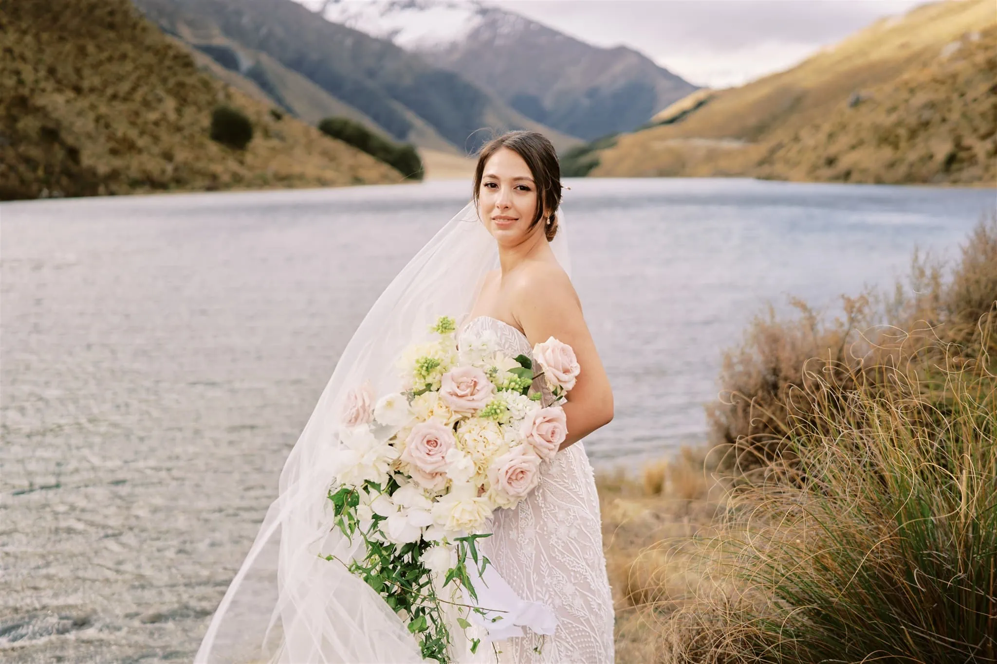 Queenstown Elopement Heli Wedding Photographer クイーンズタウン結婚式 | Mariah, the bride, standing by a lake in Queenstown with a bouquet of flowers.