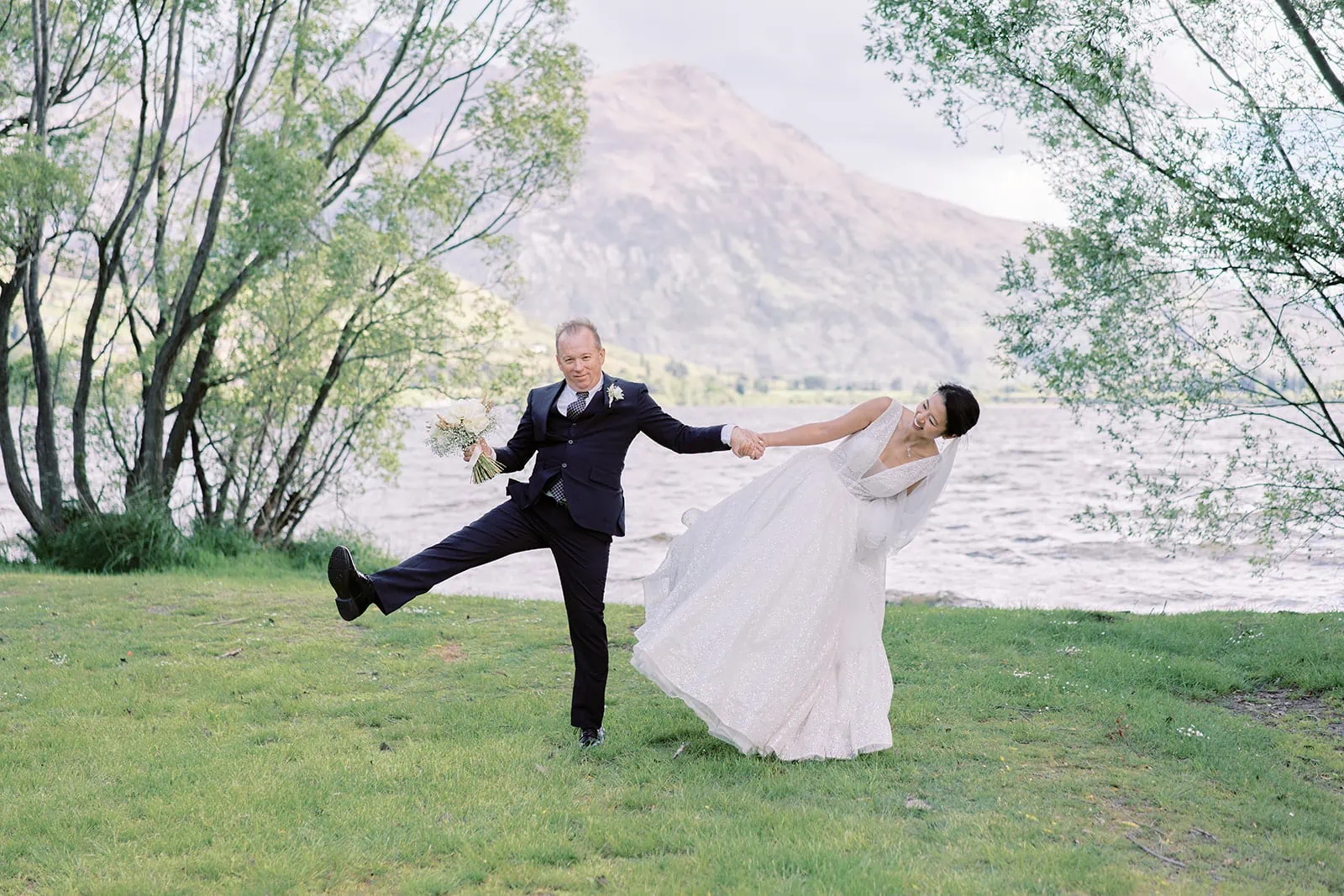 Queenstown Elopement Heli Wedding Photographer クイーンズタウン結婚式 | A bride and groom named Joel and Meng joyfully jump in the grass near Stoneridge Estate, a picturesque lake setting.