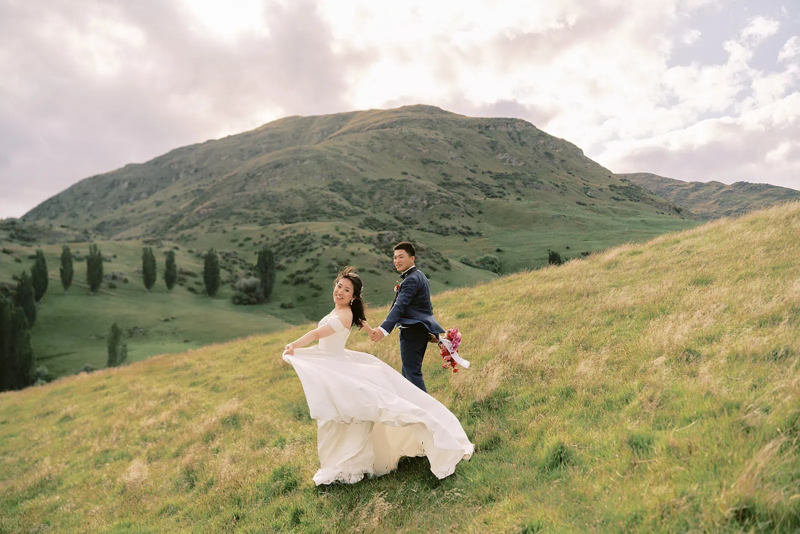 Queenstown Elopement Heli Wedding Photographer クイーンズタウン結婚式 | A couple in wedding attire joyfully running on a grassy hill during their pre-wedding photoshoot.
