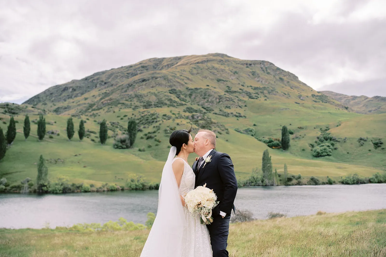 Queenstown Elopement Heli Wedding Photographer クイーンズタウン結婚式 | Joel and Meng, a bride and groom, share an affectionate kiss in front of the serene Stoneridge Estate lake.