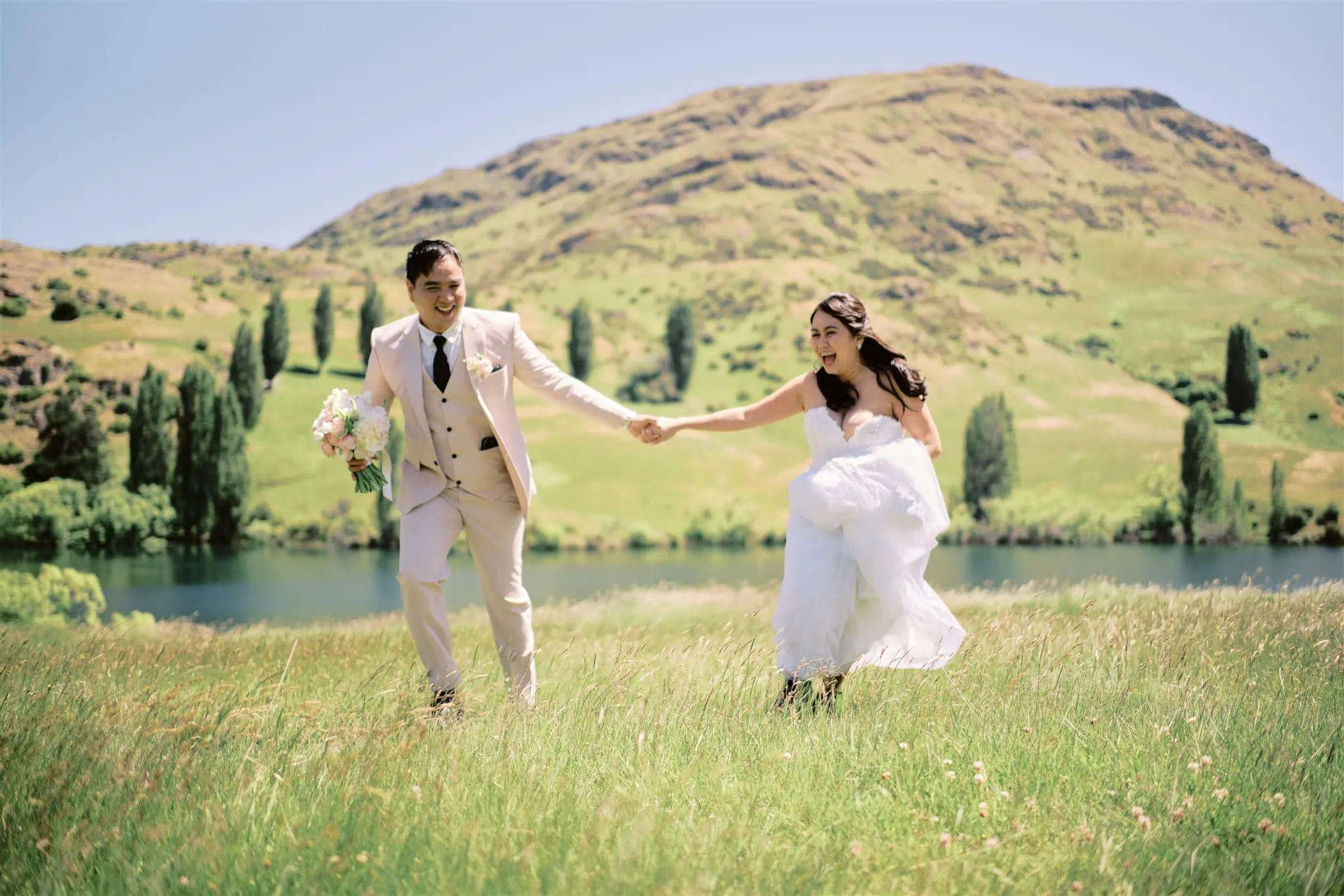 Queenstown Elopement Heli Wedding Photographer クイーンズタウン結婚式 | A couple engaged in a Queenstown elopement, holding hands and running in a field.