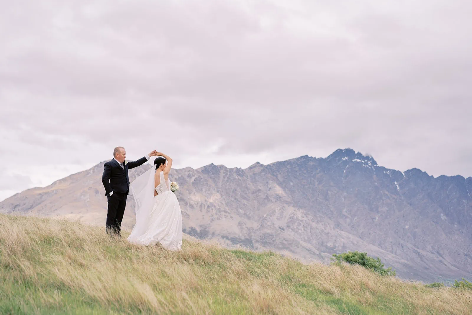 Queenstown Elopement Heli Wedding Photographer クイーンズタウン結婚式 | Meng and Joel, a bride and groom, standing on a grassy hill with mountains in the background at Stoneridge Estate.