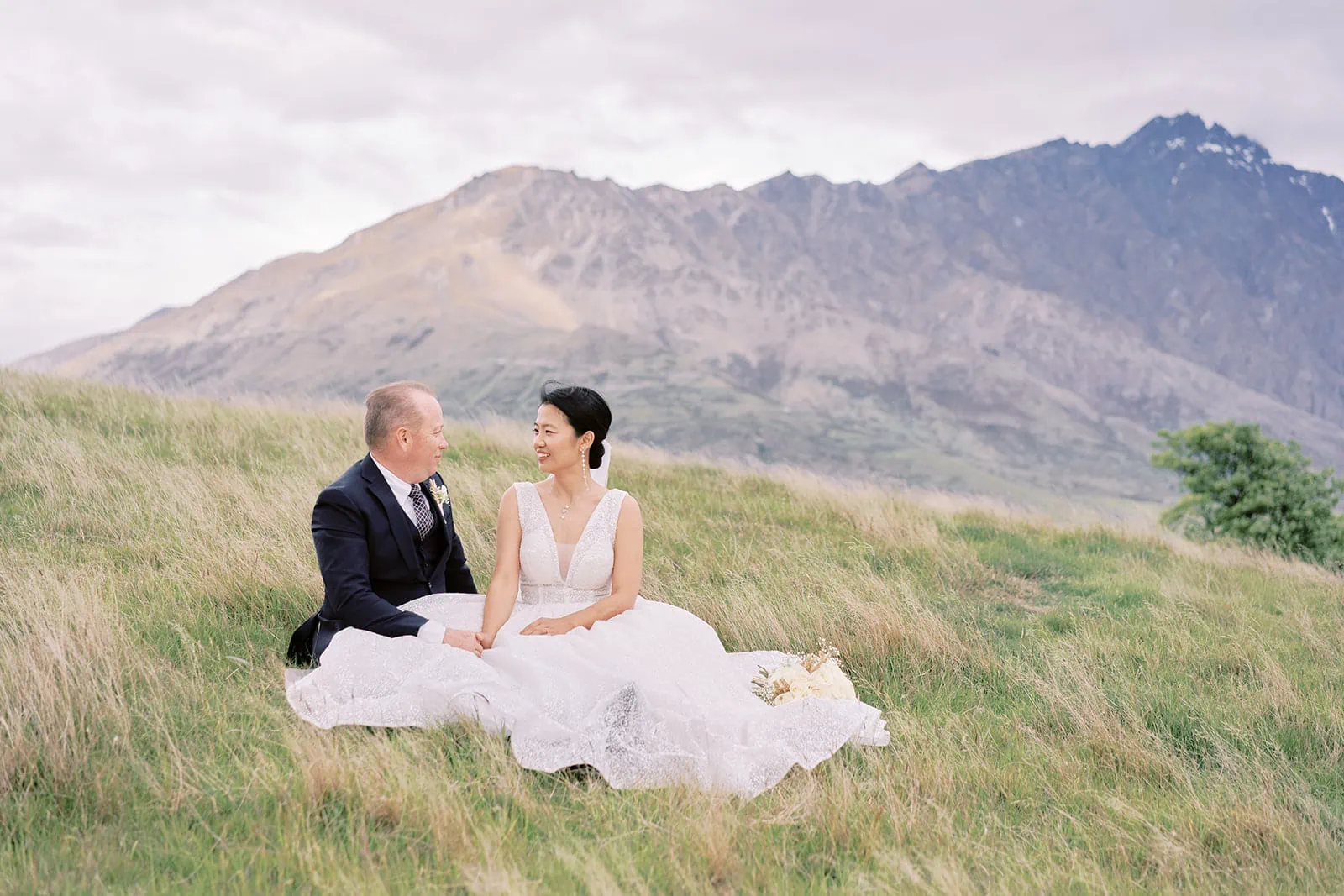 Queenstown Elopement Heli Wedding Photographer クイーンズタウン結婚式 | Joel and Meng sitting in grass with mountains in the background at Stoneridge Estate.
