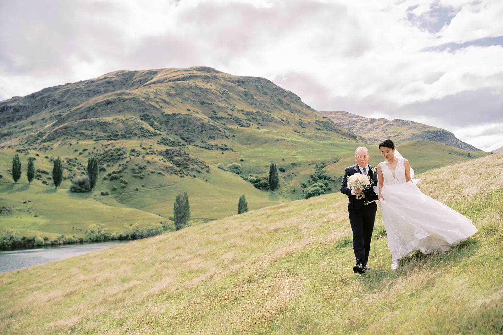 Queenstown Elopement Heli Wedding Photographer クイーンズタウン結婚式 | Meng & Joel's Stoneridge Estate Wedding captures the couple's romantic stroll through a captivating grassy field, surrounded by majestic mountains in the background.