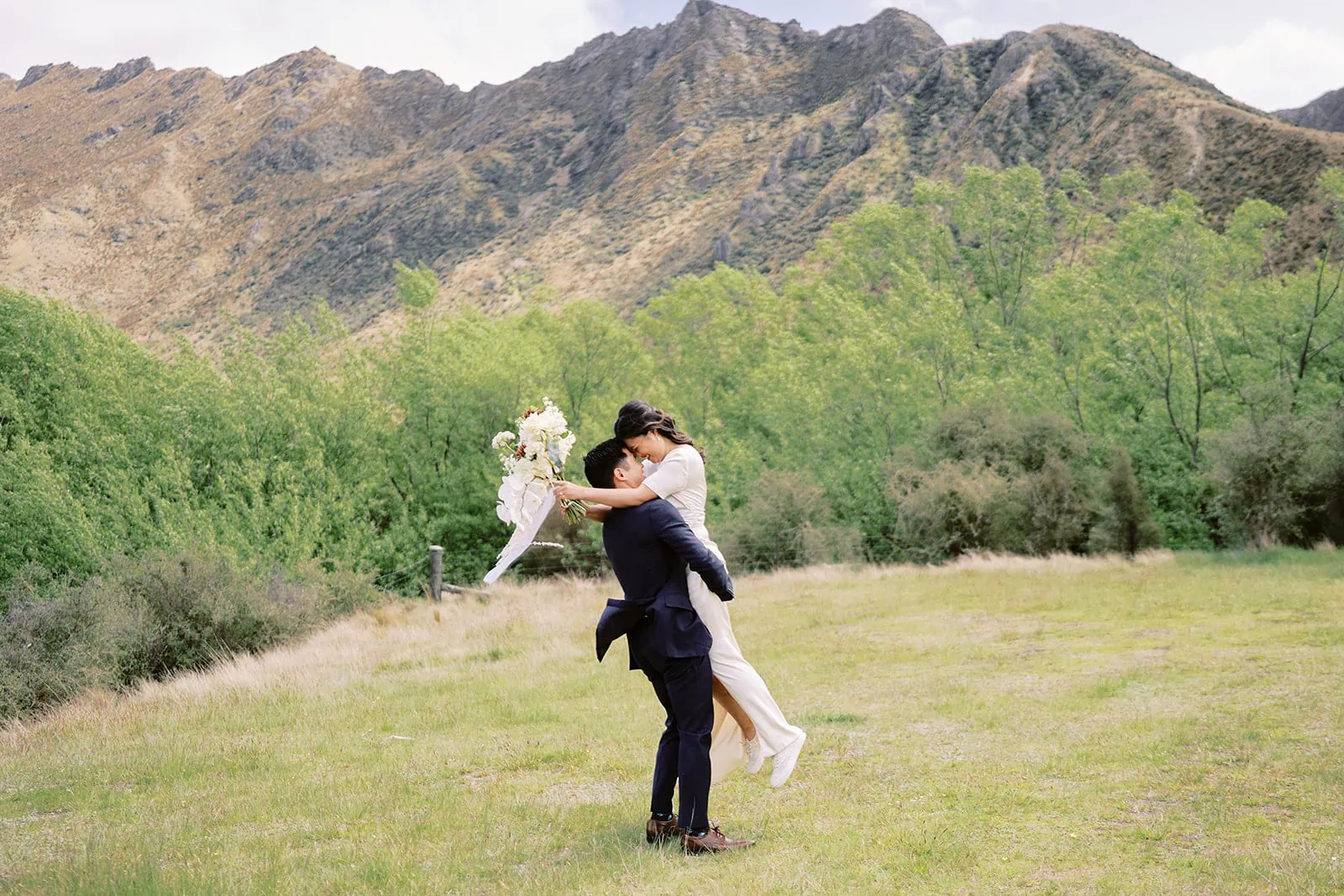 Queenstown Elopement Heli Wedding Photographer クイーンズタウン結婚式 | A pre-wedding photoshoot capturing a romantic moment as a man and woman passionately kiss in a stunning field with majestic mountains in the background.