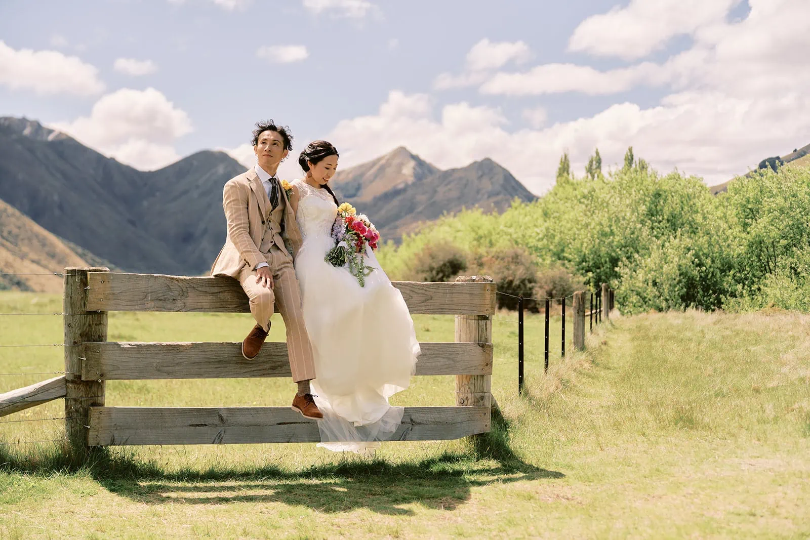 Queenstown Elopement Heli Wedding Photographer クイーンズタウン結婚式 | A bride and groom sitting on a fence overlooking the picturesque mountains of Queenstown.