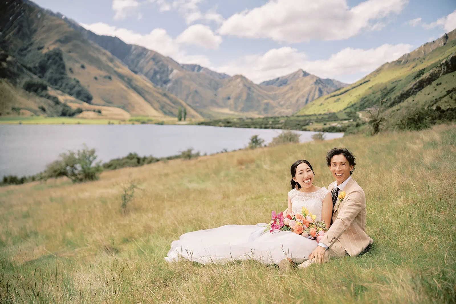 Queenstown Elopement Heli Wedding Photographer クイーンズタウン結婚式 | A pre-wedding couple sitting on a grassy field near a lake in Queenstown.