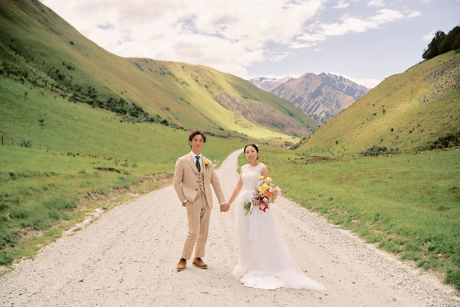 Queenstown Elopement Heli Wedding Photographer クイーンズタウン結婚式 | A bride and groom standing on a dirt road in Queenstown, surrounded by majestic mountains.