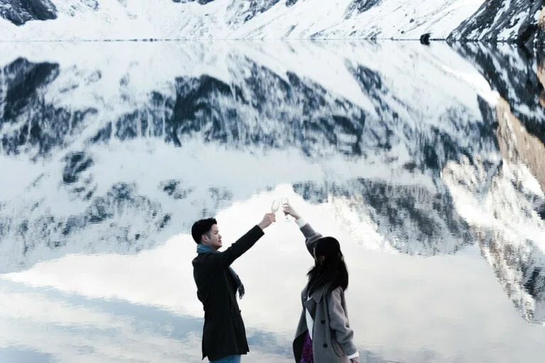 Queenstown Elopement Heli Wedding Photographer クイーンズタウン結婚式 | Dee and Hiroki, a couple, holding a champagne glass in front of a snow-covered lake near Queenstown.