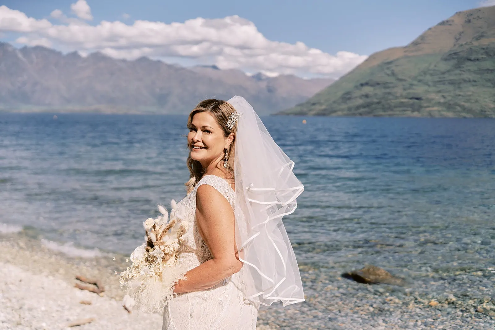 Queenstown Elopement Heli Wedding Photographer クイーンズタウン結婚式 | Melissa, a bride, standing on the shore of Lake Wanaka with mountains in the background.