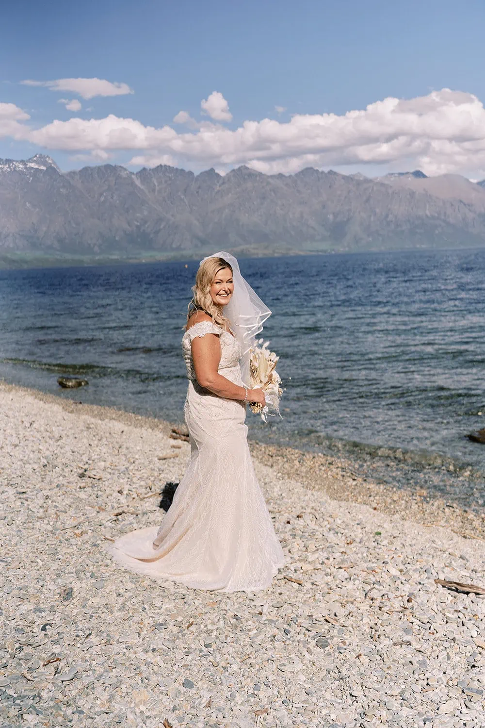 Queenstown Elopement Heli Wedding Photographer クイーンズタウン結婚式 | Melissa, a bride, standing on a beach with mountains in the background at Kamana Lakehouse Queenstown for her elopement with Scott.