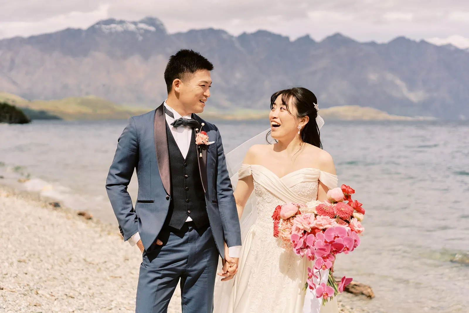 Queenstown Elopement Heli Wedding Photographer クイーンズタウン結婚式 | A couple dressed in their wedding attire pose for a pre-wedding photoshoot on a picturesque beach, with majestic mountains towering in the background.