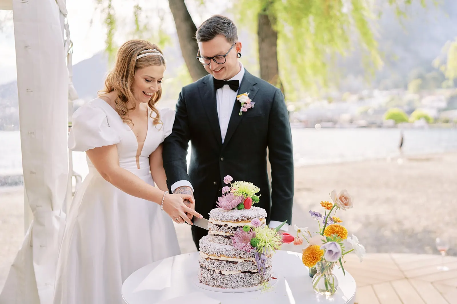 Queenstown Elopement Heli Wedding Photographer クイーンズタウン結婚式 | A bride and groom cutting their wedding cake in front of a lake in Queenstown.