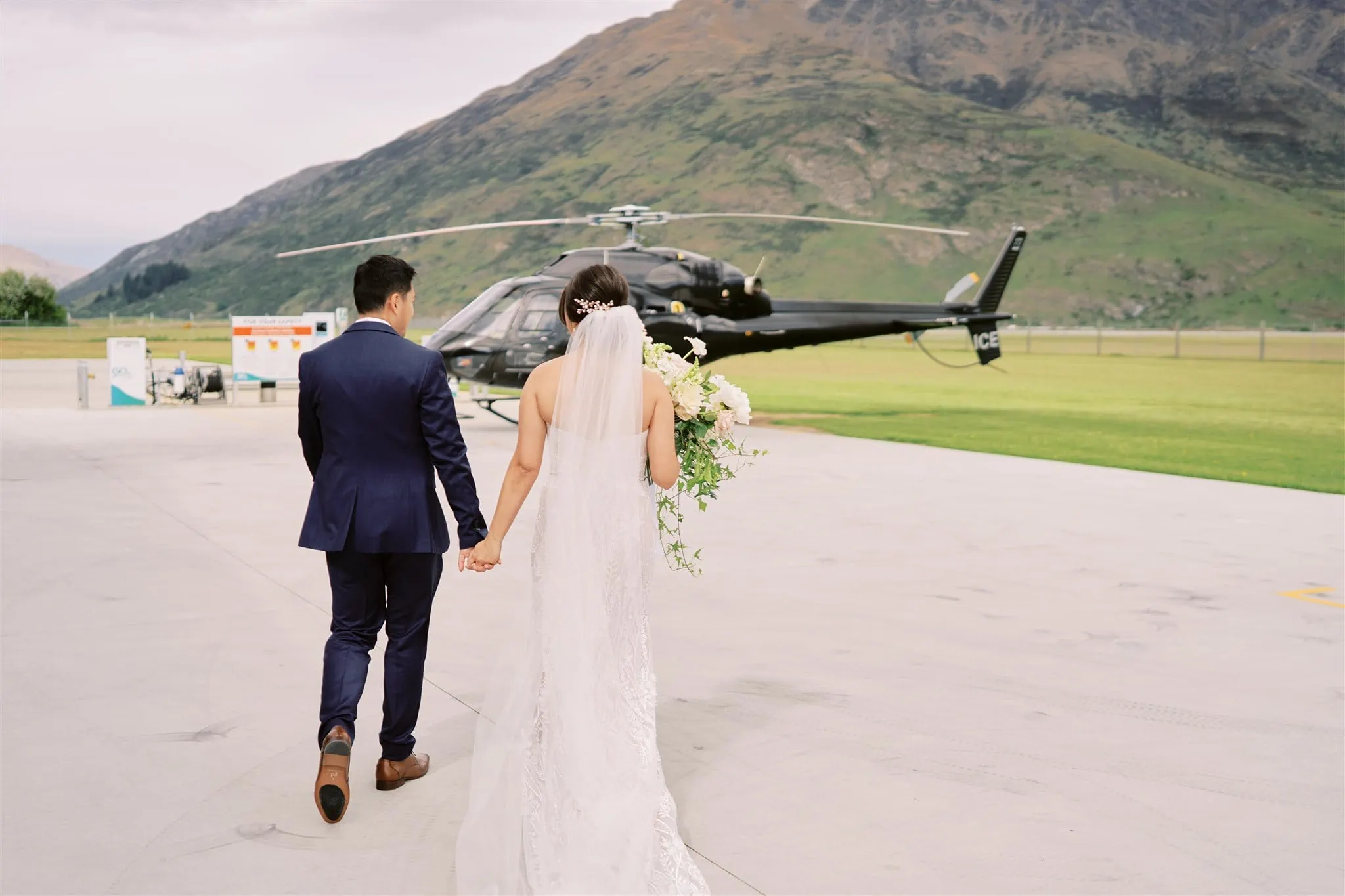 Queenstown Elopement Heli Wedding Photographer クイーンズタウン結婚式 | A bride and groom walk past a helicopter in front of mountains in Queenstown.