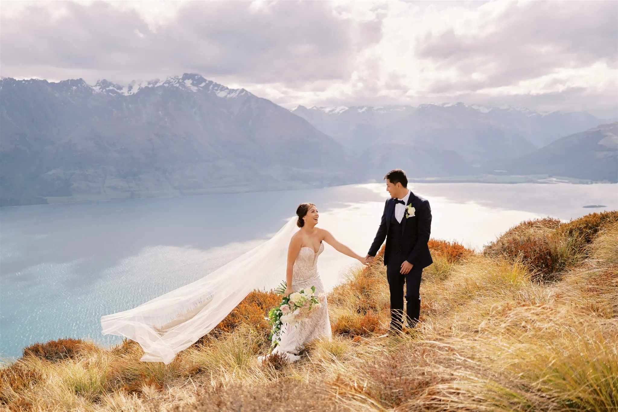 Queenstown Elopement Heli Wedding Photographer クイーンズタウン結婚式 | Mariah, the bride, and her groom standing on top of a cliff overlooking lake Wanaka in an epic Queenstown setting.