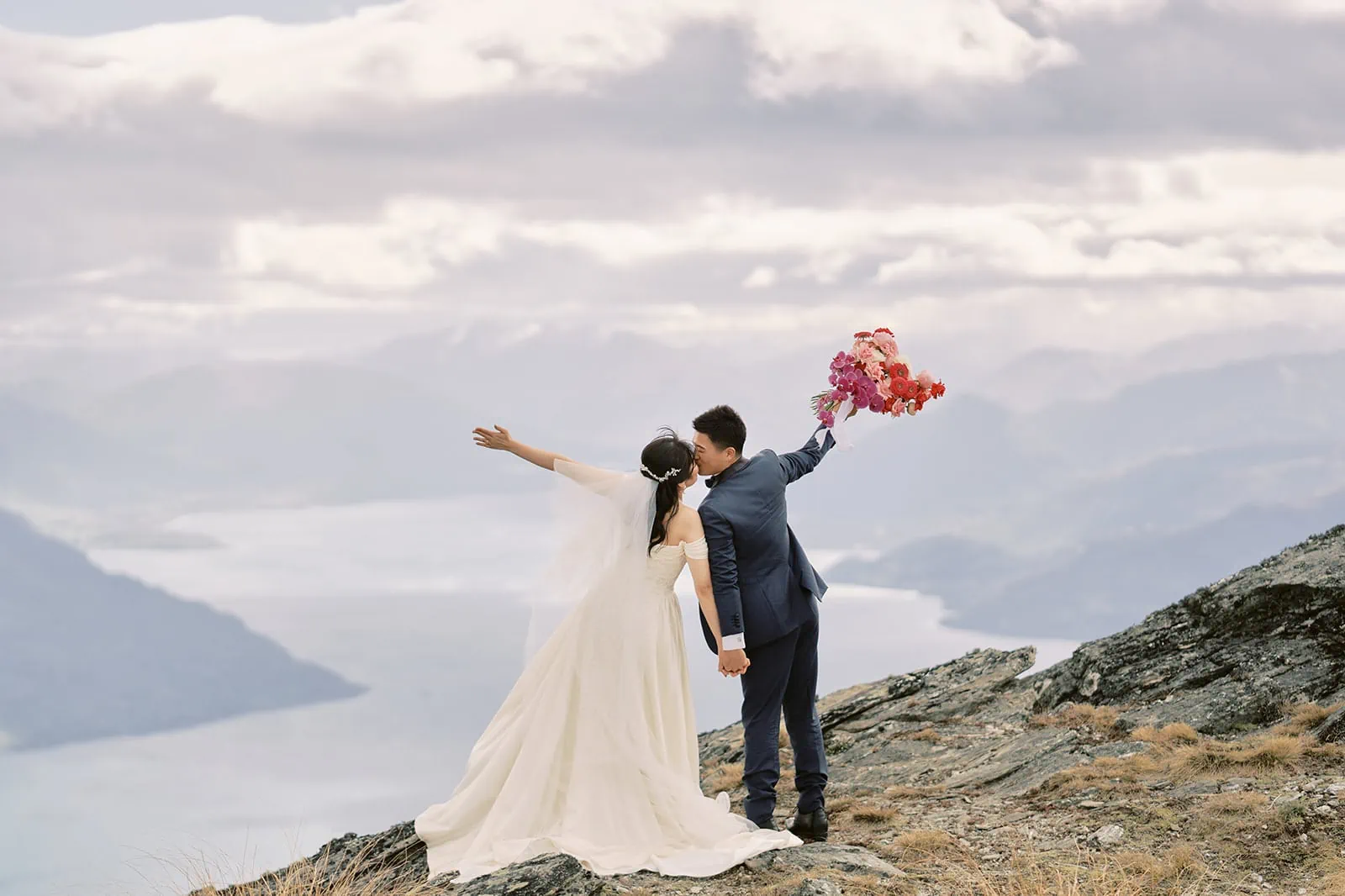 Queenstown Elopement Heli Wedding Photographer クイーンズタウン結婚式 | A couple, dressed in wedding attire, poses for a pre-wedding photoshoot on top of a mountain overlooking Lake Wanaka.