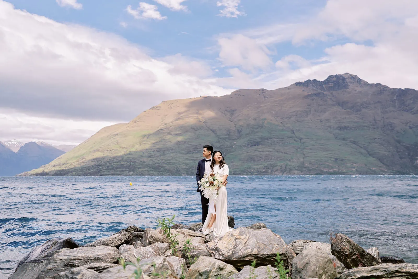 Queenstown Elopement Heli Wedding Photographer クイーンズタウン結婚式 | A romantic pre-wedding photoshoot of a man and woman standing on rocks by water and mountains.