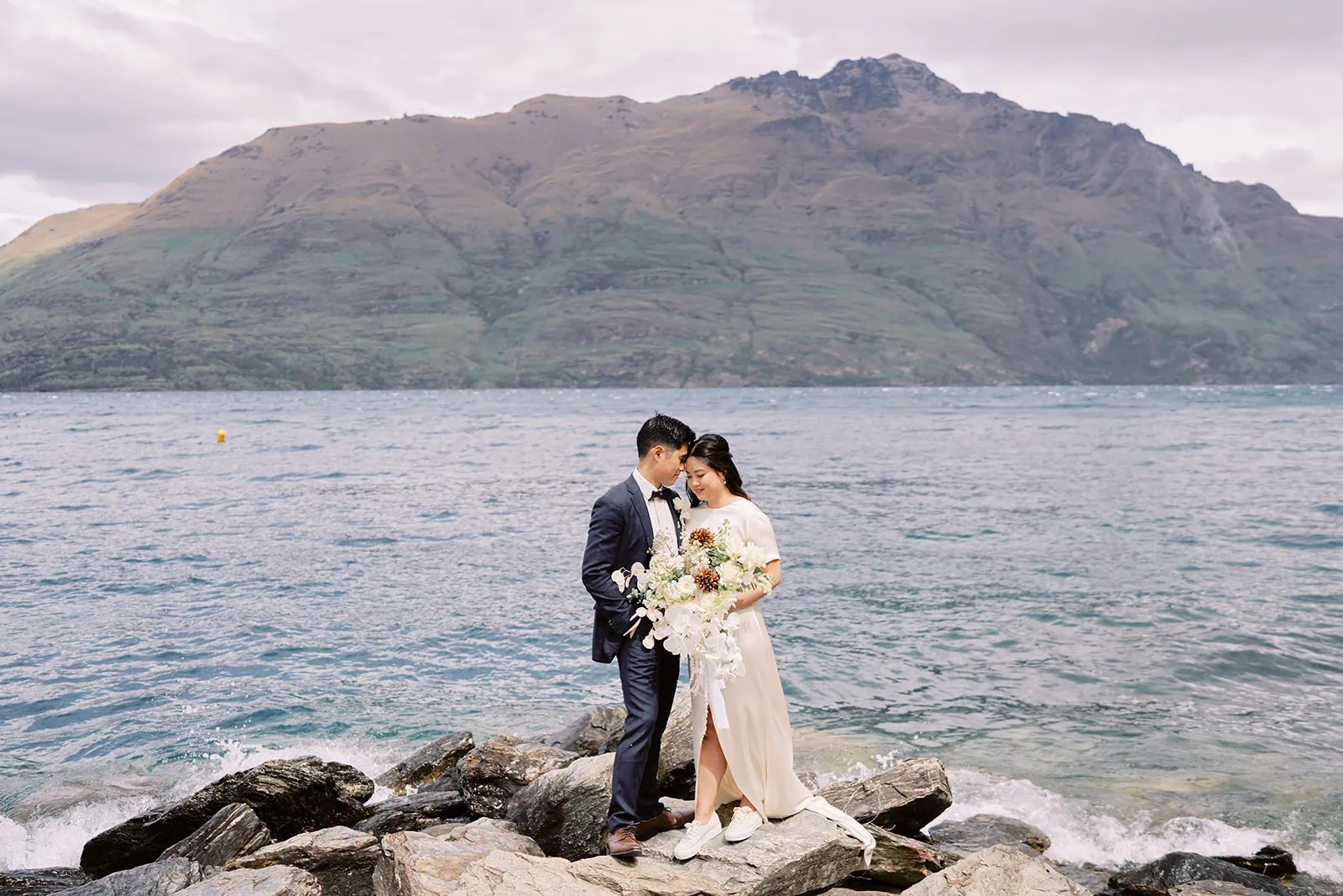 Queenstown Elopement Heli Wedding Photographer クイーンズタウン結婚式 | A man and woman having a pre-wedding photoshoot, standing on rocks by water with mountains in the background.