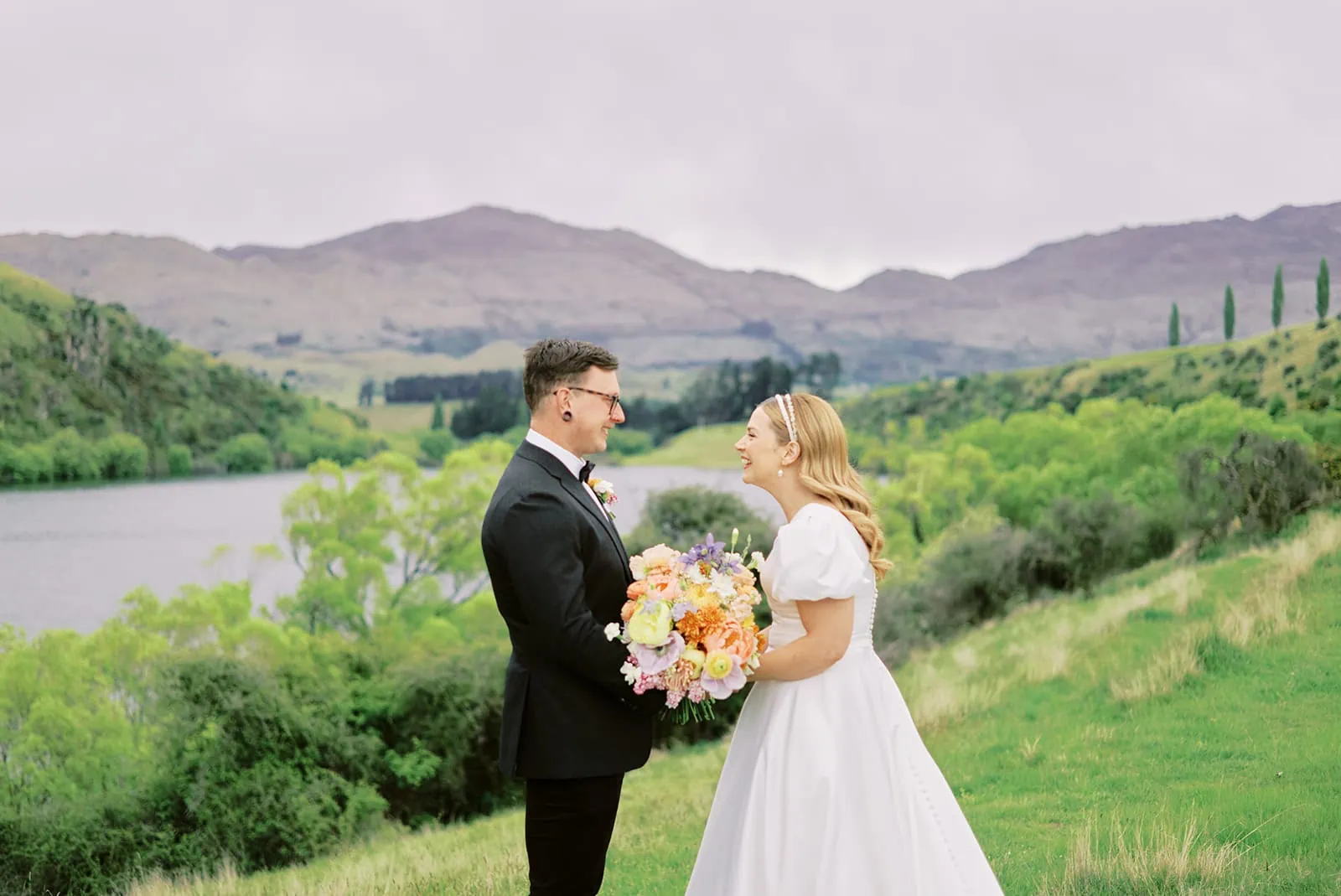 Queenstown New Zealand Heli Wedding Elopement Photographer クイーンズタウン　ニュージーランド　エロープメント 結婚式 | A couple in wedding attire smiling at each other outdoors with a bouquet and Lake Johnson in the background.
