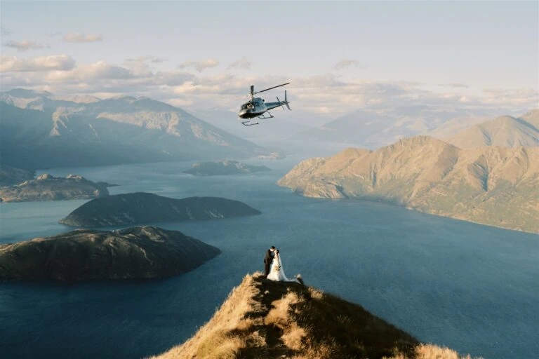 Queenstown New Zealand Heli Wedding Elopement Photographer クイーンズタウン　ニュージーランド　エロープメント 結婚式 | Betty & Nick stand on a mountain peak overlooking Moke Lake with a helicopter flying in the background during their Queenstown elopement.