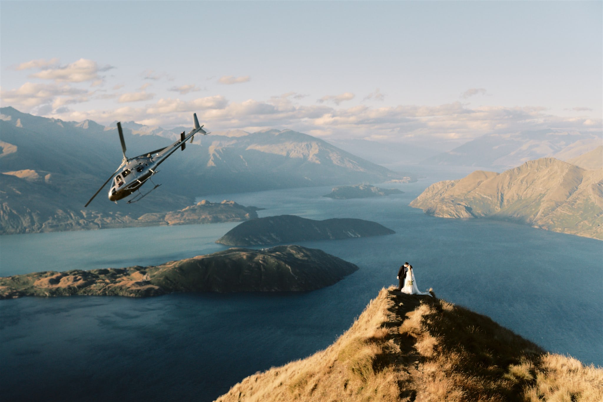 Queenstown New Zealand Heli Wedding Elopement Photographer クイーンズタウン ニュージーランド エロープメント 結婚式 | A couple, Betty & Nick, stands on a mountain ridge overlooking Moke Lake as a helicopter flies nearby during their Queenstown elopement.
