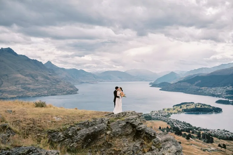 Queenstown Tekapo New Zealand Heli Wedding Elopement Pre-Wedding Shoot Photographer クイーンズタウン　テカポ　ニュージーランド　エロープメント 前撮り　フォトウェディング　結婚式 | Aoi & Conor embrace on a rocky overlook with a vast lake and mountains in the background during their Queenstown pre-wedding shoot.