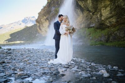 Queenstown New Zealand Heli Wedding Elopement Photographer クイーンズタウン　ニュージーランド　エロープメント 結婚式 | A couple in wedding attire standing by a river with a waterfall in the background, celebrating their elopement wedding.