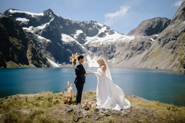 Queenstown New Zealand Heli Wedding Elopement Photographer クイーンズタウン　ニュージーランド　エロープメント 結婚式 | An elopement wedding couple standing by a mountain lake with a scenic backdrop of snow-capped peaks.