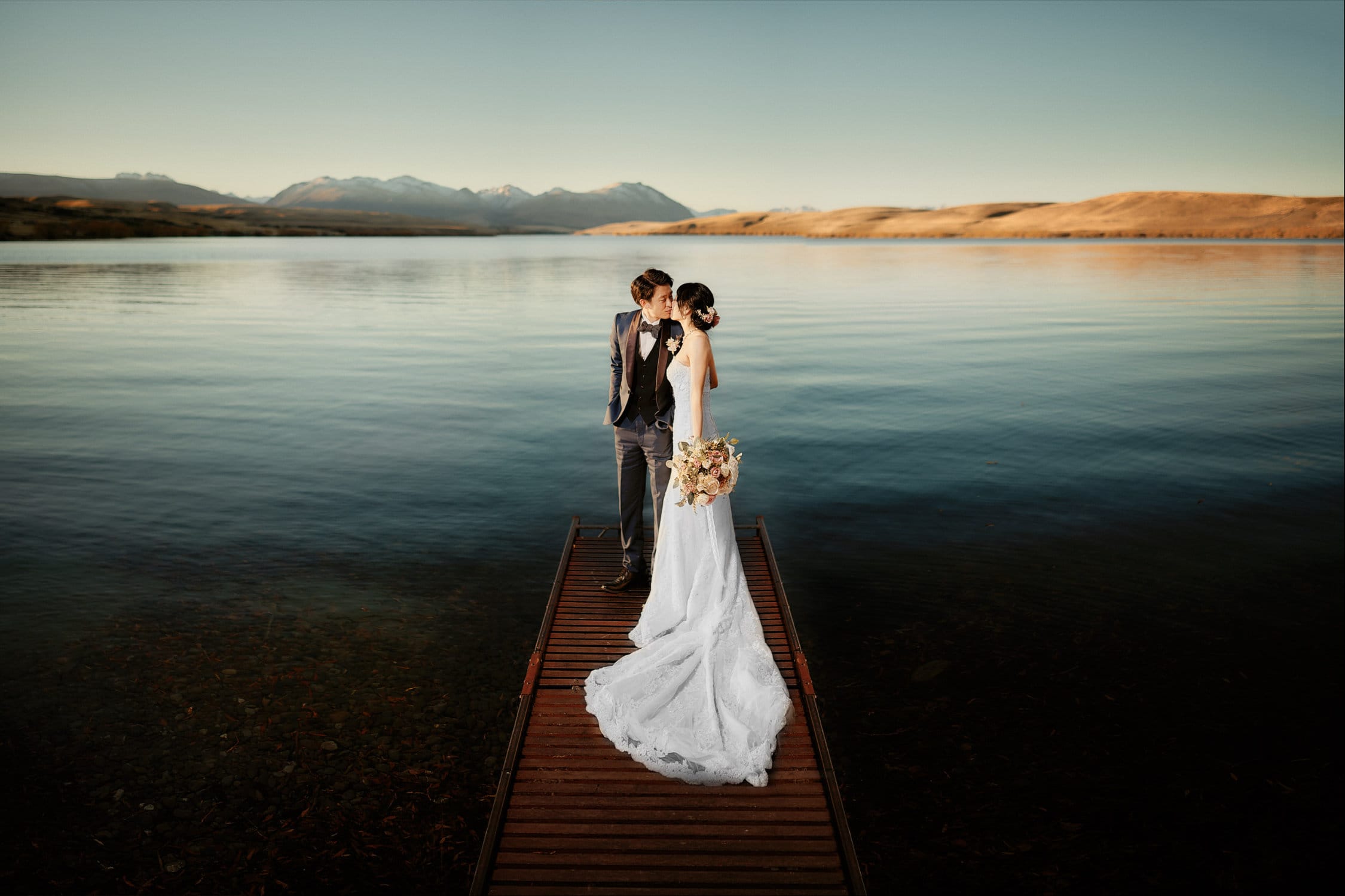 Queenstown Elopement Heli Wedding Photographer クイーンズタウン結婚式 | A bride and groom standing on a dock in front of a lake.