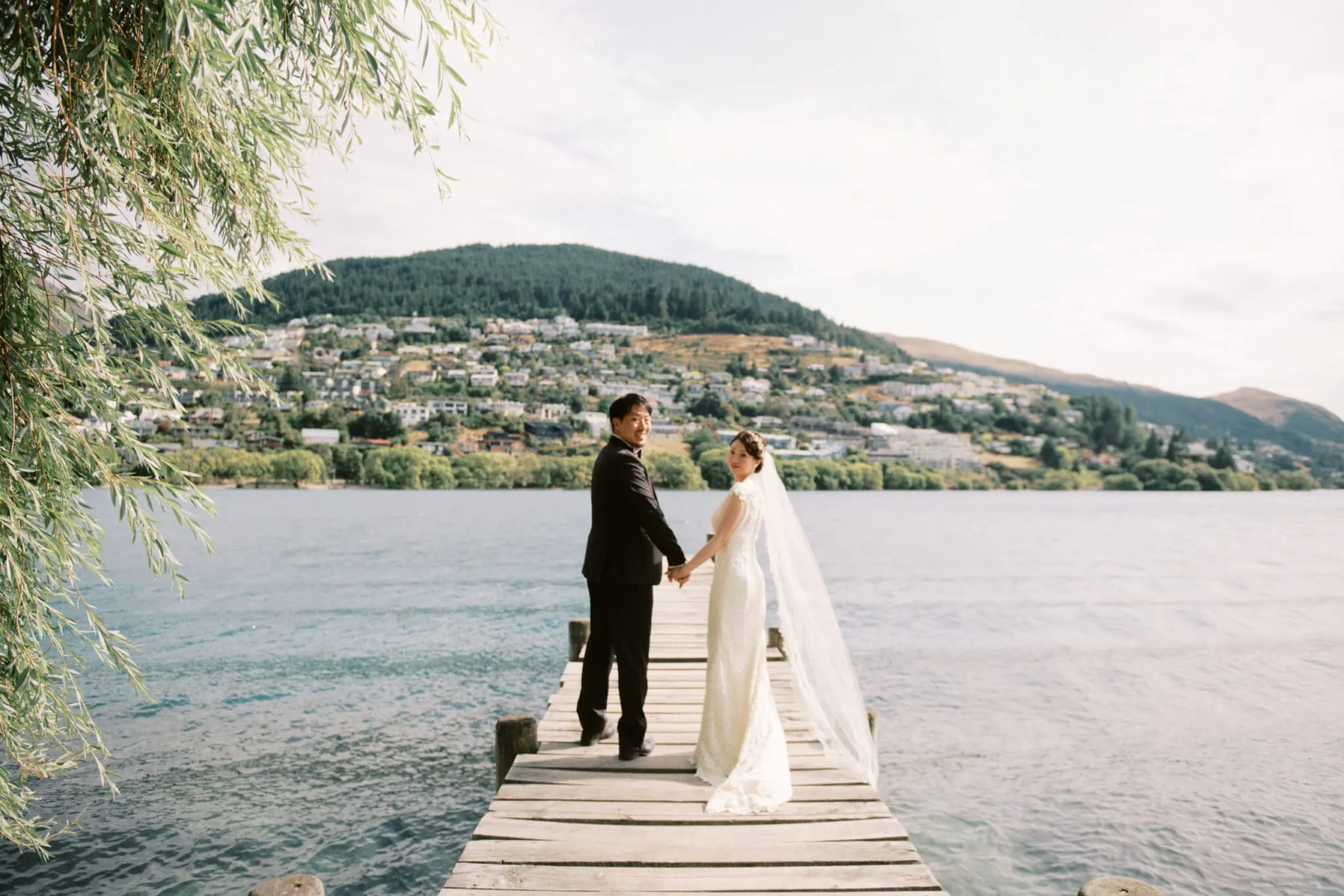 Queenstown Tekapo New Zealand Heli Wedding Elopement Pre-Wedding Shoot Photographer クイーンズタウン　テカポ　ニュージーランド　エロープメント 前撮り　フォトウェディング　結婚式 | Yuko & Shuntaro hold hands on a wooden dock overlooking a serene lake with a scenic hillside town in the background during their Tekapo Pre-Wedding Shoot.