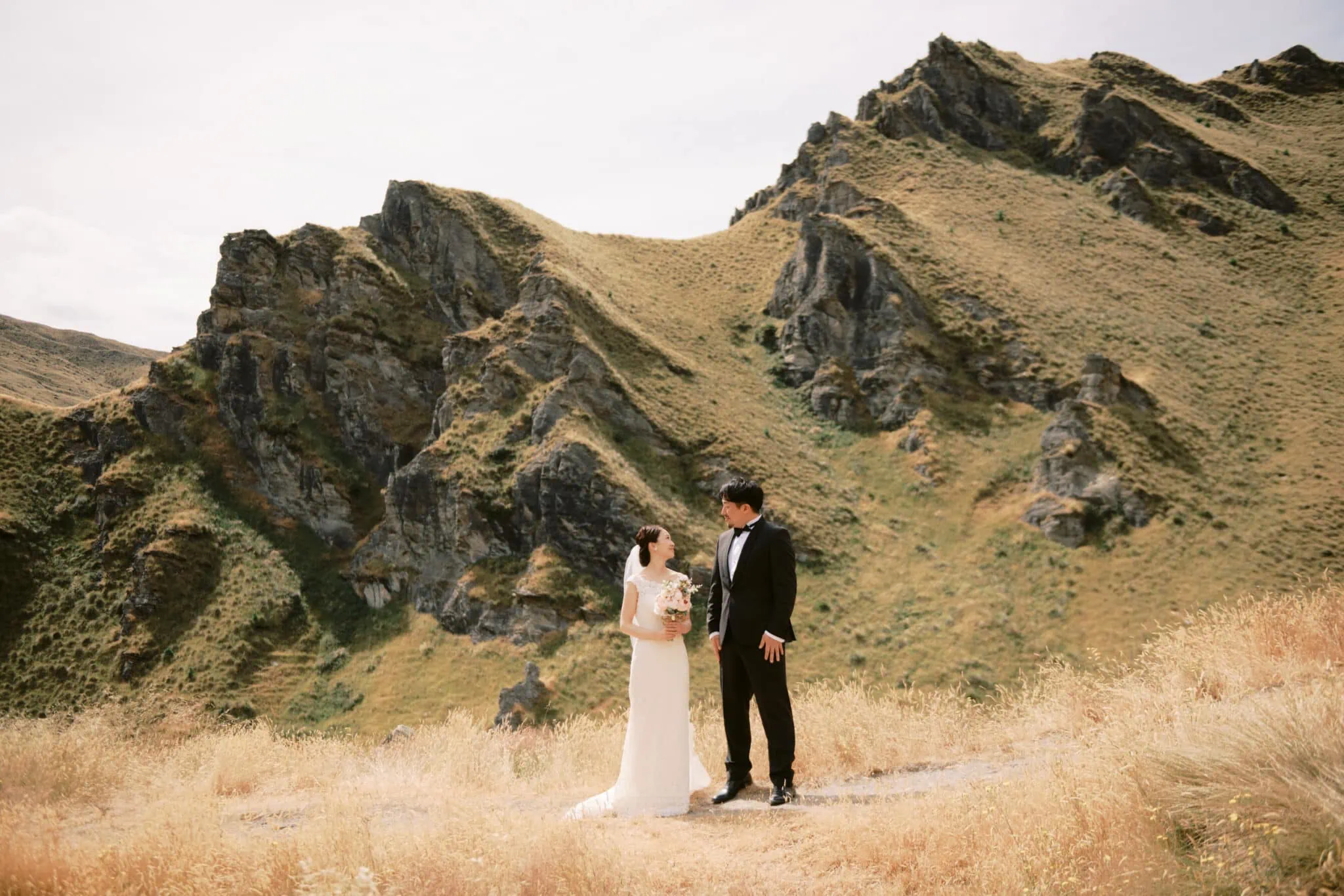 Queenstown Tekapo New Zealand Heli Wedding Elopement Pre-Wedding Shoot Photographer クイーンズタウン　テカポ　ニュージーランド　エロープメント 前撮り　フォトウェディング　結婚式 | Yuko & Shuntaro, in wedding attire, holding hands in a scenic mountainous landscape during their Tekapo Pre-Wedding Shoot.