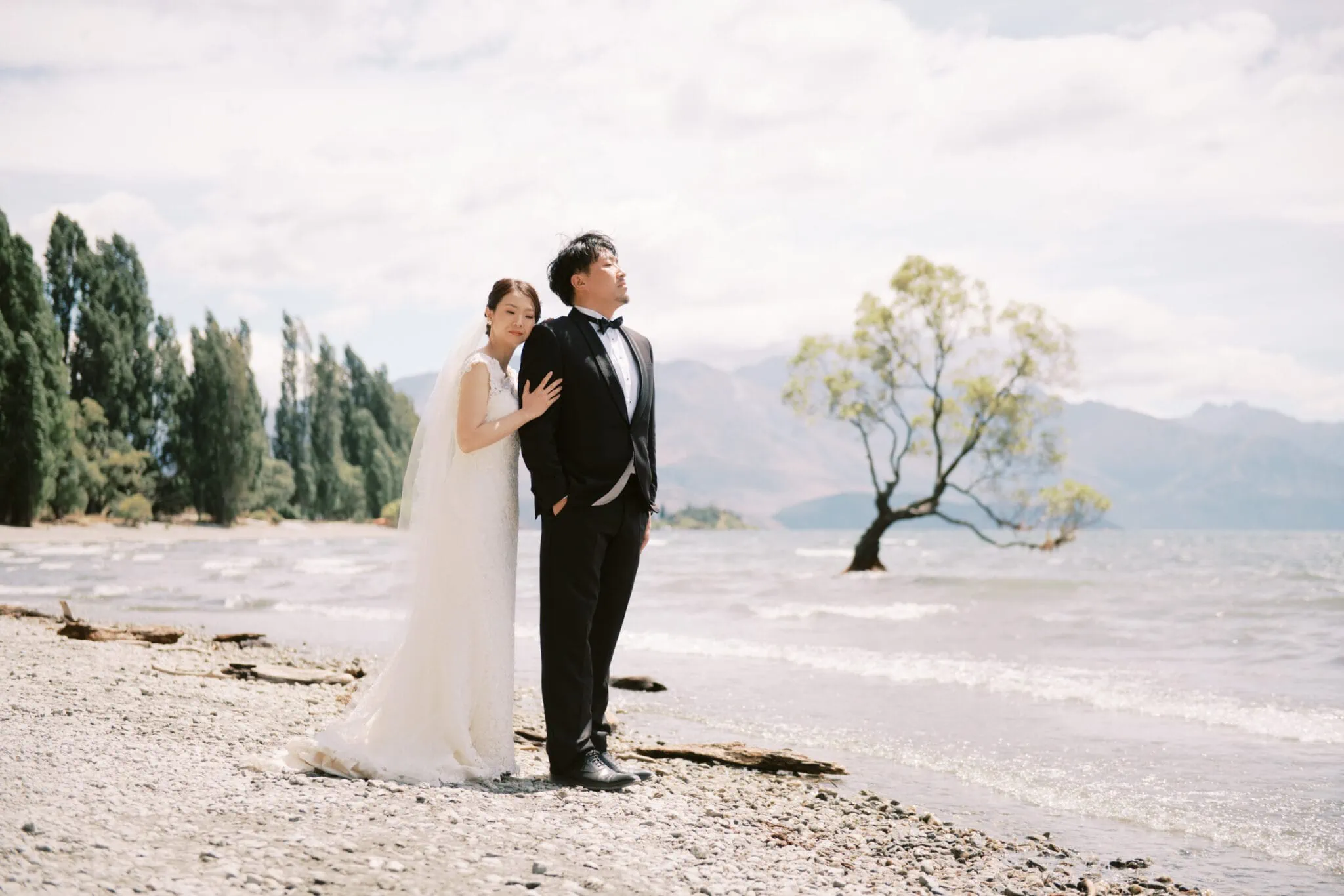 Queenstown Tekapo New Zealand Heli Wedding Elopement Pre-Wedding Shoot Photographer クイーンズタウン　テカポ　ニュージーランド　エロープメント 前撮り　フォトウェディング　結婚式 | Yuko & Shuntaro standing together on a beach with trees and a lake in the background during their Tekapo Pre-Wedding Shoot.