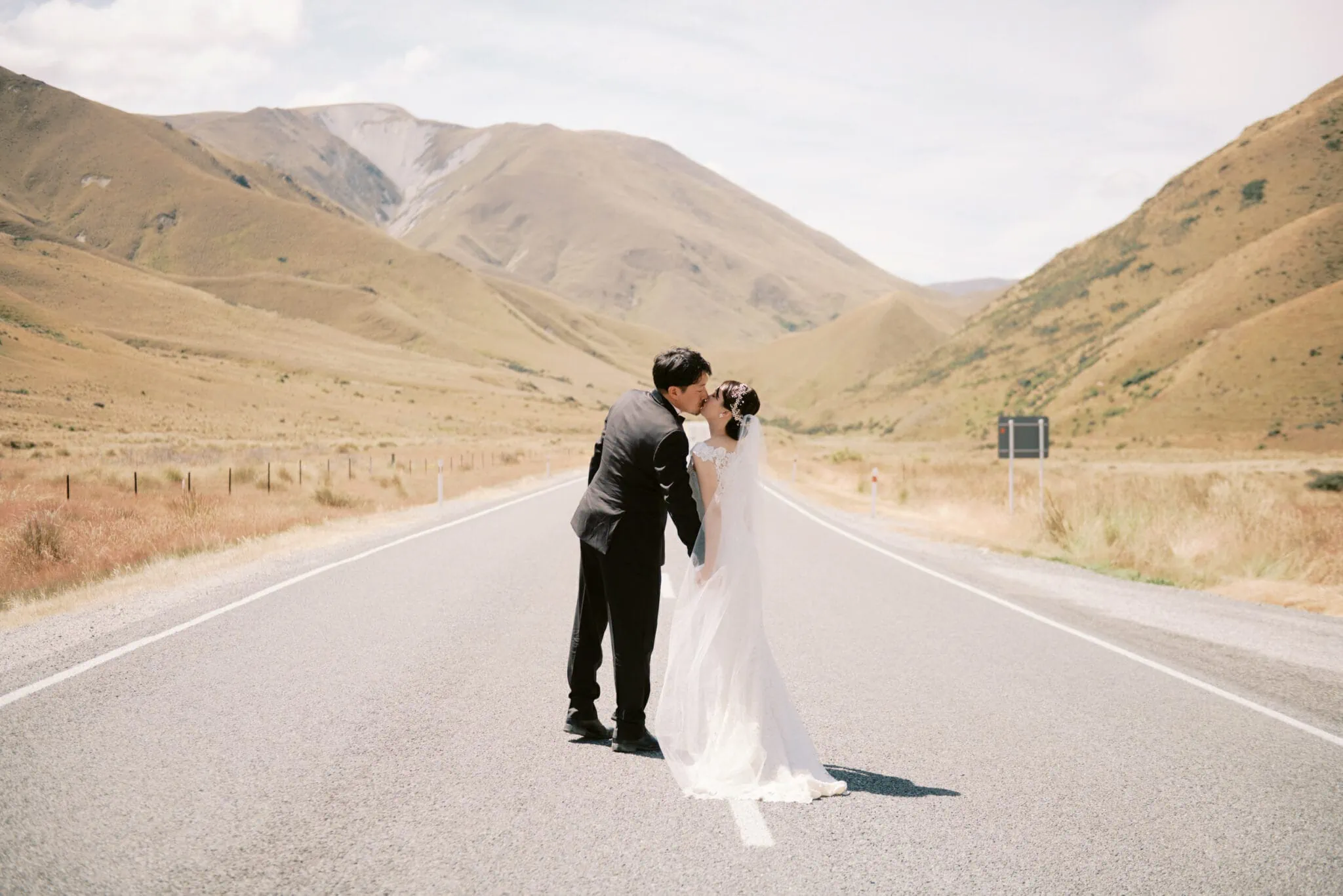 Queenstown Tekapo New Zealand Heli Wedding Elopement Pre-Wedding Shoot Photographer クイーンズタウン　テカポ　ニュージーランド　エロープメント 前撮り　フォトウェディング　結婚式 | Bride and groom sharing a kiss in the middle of a road with mountainous landscape in the background during their Tekapo Pre-Wedding Shoot.