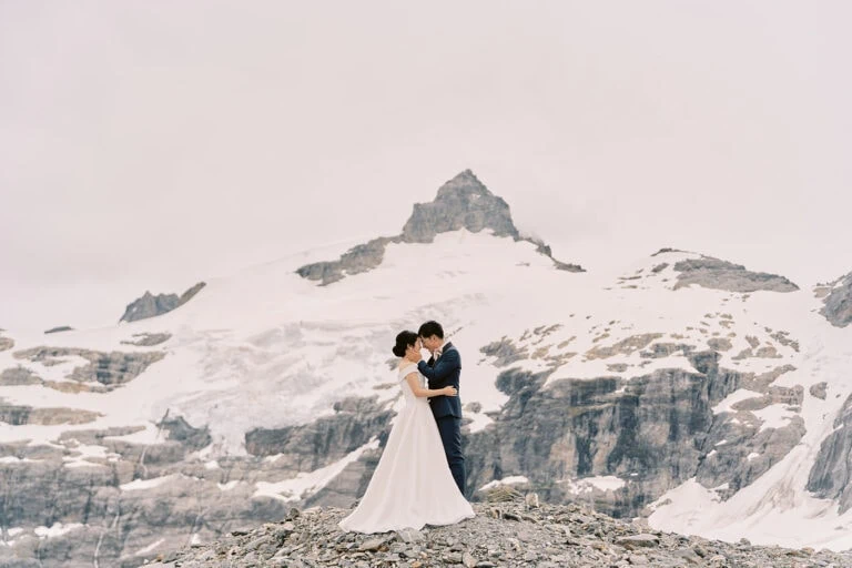 Queenstown Elopement Heli Wedding Photographer クイーンズタウン結婚式 | A man and Sakiko in a white dress kissing in front of a snowy mountain during their Queenstown Heli Elopement.