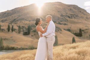 クイーンズタウン　テカポ　ニュージーランド　エロープメント 前撮り　フォトウェディング　結婚式 | クイーンズタウンでの結婚式で、フォーマルな服装をしたカップルが、日没の山を背景にした太陽の光が降り注ぐ野原で愛情深く抱き合っています。