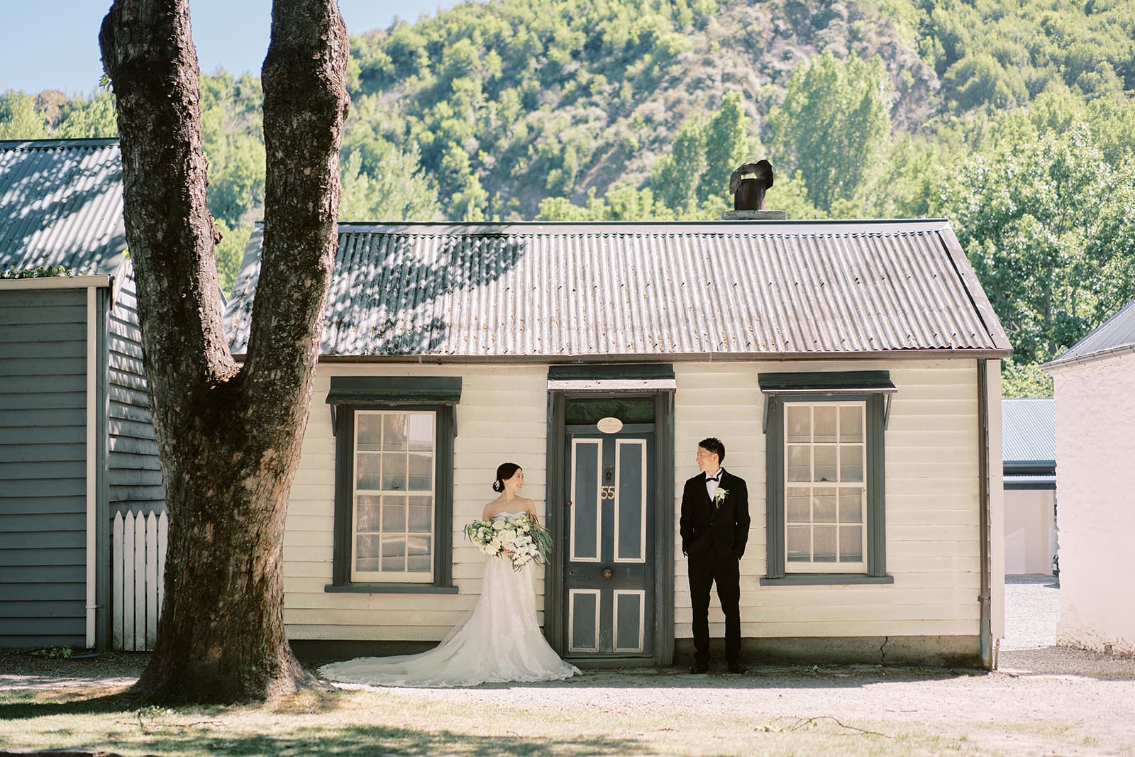 クイーンズタウン　テカポ　ニュージーランド　エロープメント 前撮り　フォトウェディング　結婚式 | 晴れた日に、ニュージーランドの木々に囲まれた趣のある木造コテージの外に立つ新郎新婦。