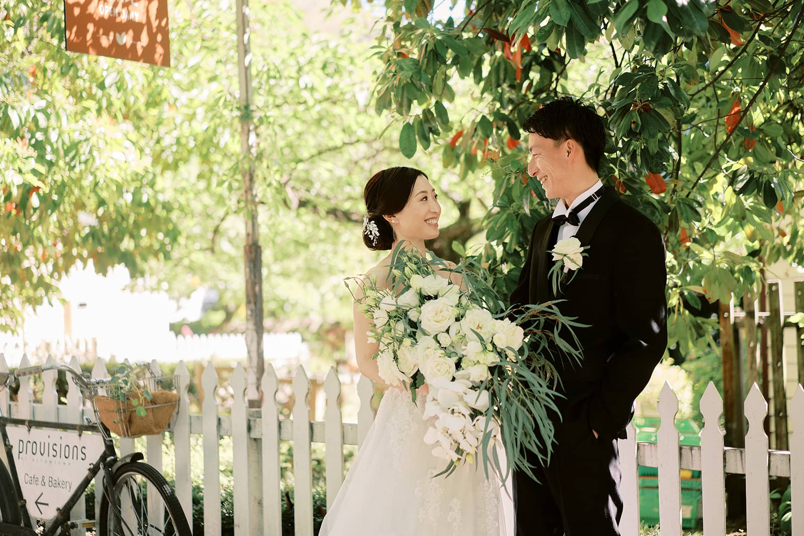 クイーンズタウン　テカポ　ニュージーランド　エロープメント 前撮り　フォトウェディング　結婚式 | 屋外に立って木々に囲まれ、ブーケを持ちながら互いに微笑み合う新郎新婦。近くには自転車と「食料」と書かれた看板がある。