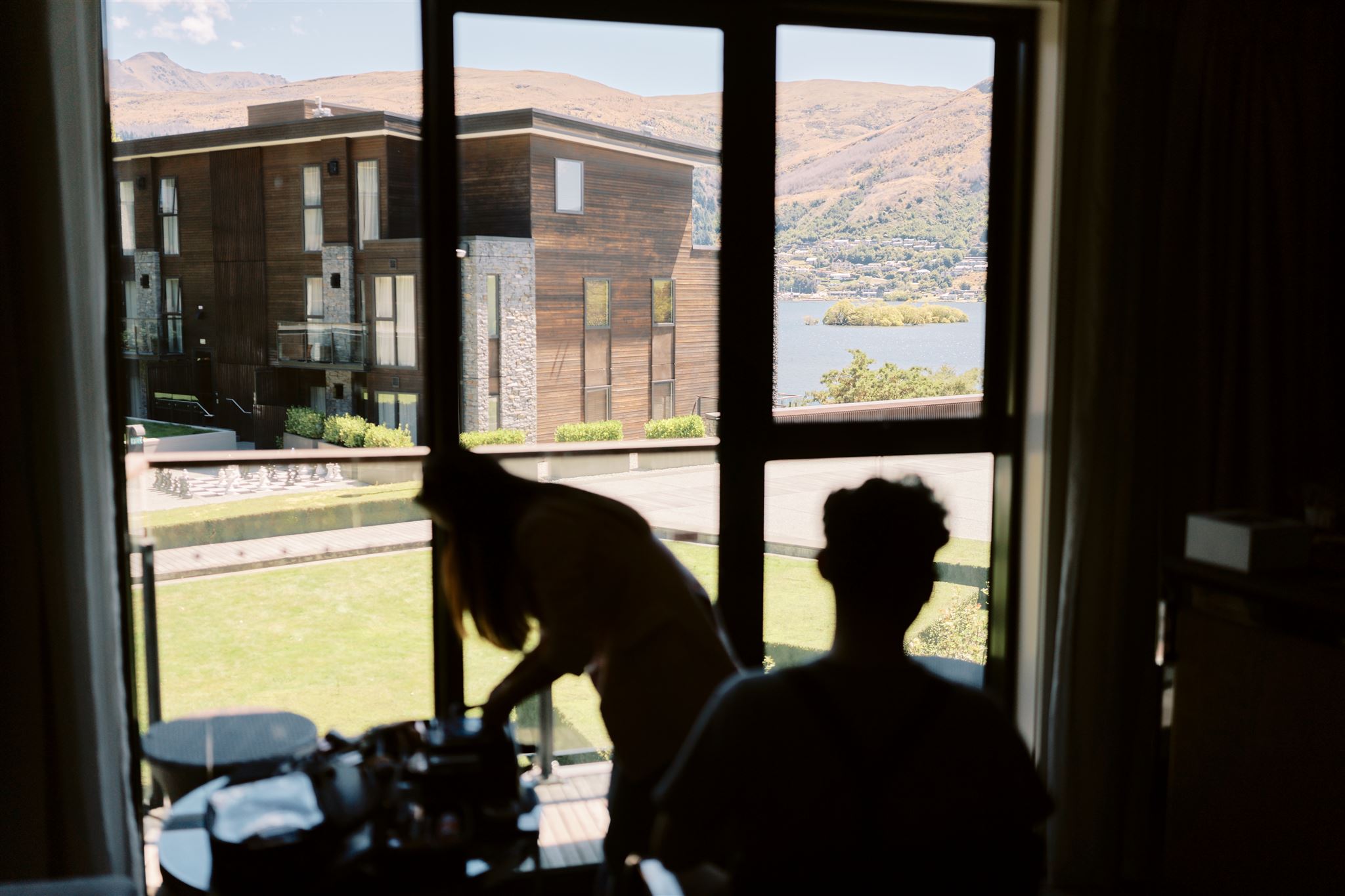 クイーンズタウン　テカポ　ニュージーランド　エロープメント 前撮り　フォトウェディング　結婚式 | 晴れた日のクイーンズタウンにある静かな湖と家々を見渡す窓に向かって、1 人は立って、もう 1 人は座っている、屋内の 2 人のシルエット。