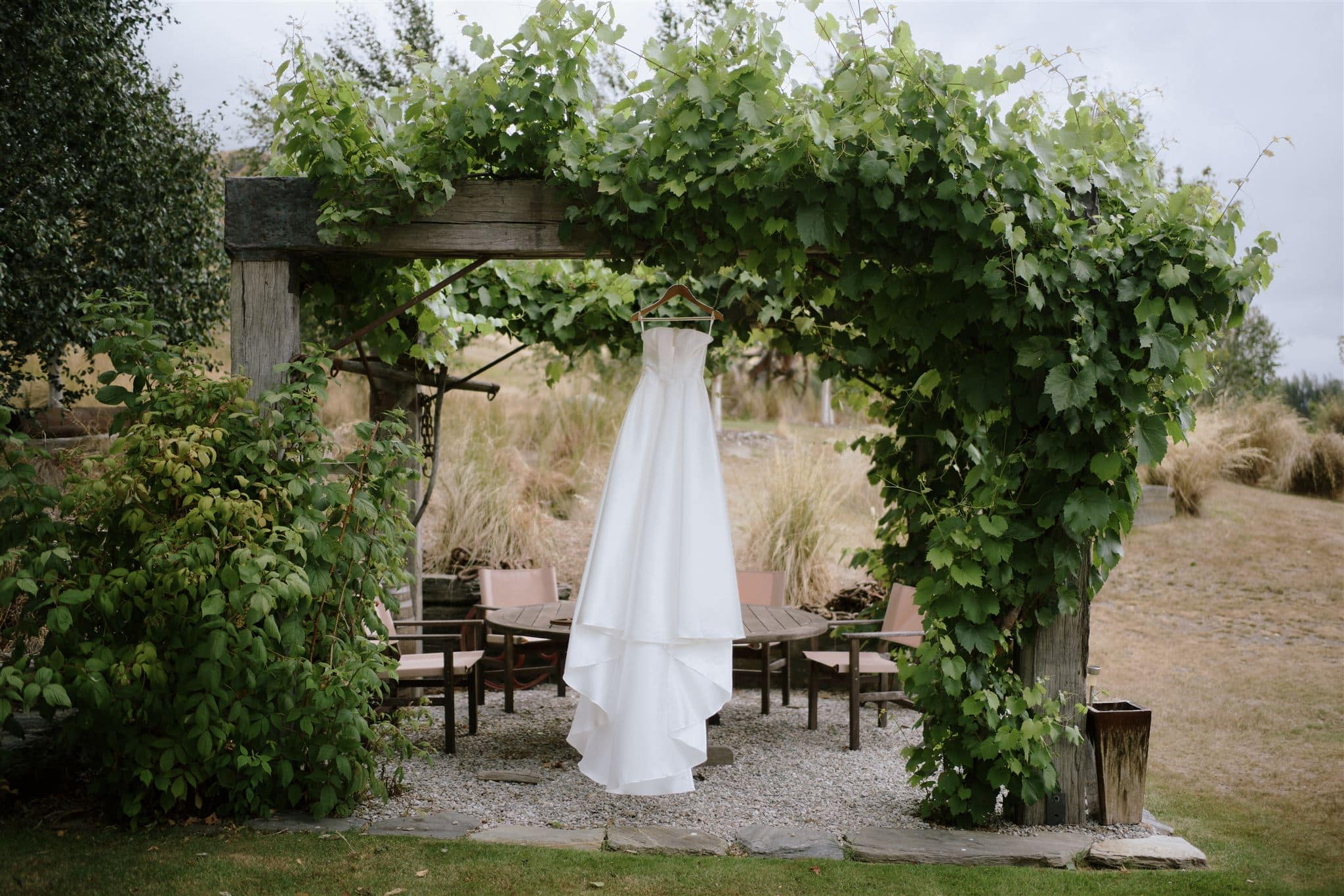 クイーンズタウン　テカポ　ニュージーランド　エロープメント 前撮り　フォトウェディング　結婚式 | ニュージーランドのクイーンズタウンの美しい景色の中、緑豊かなつる植物で覆われた木製のトレリスにウェディングドレスが吊るされており、その下には素朴なダイニングが設えられた静かな屋外ガーデンエリアが広がっています。