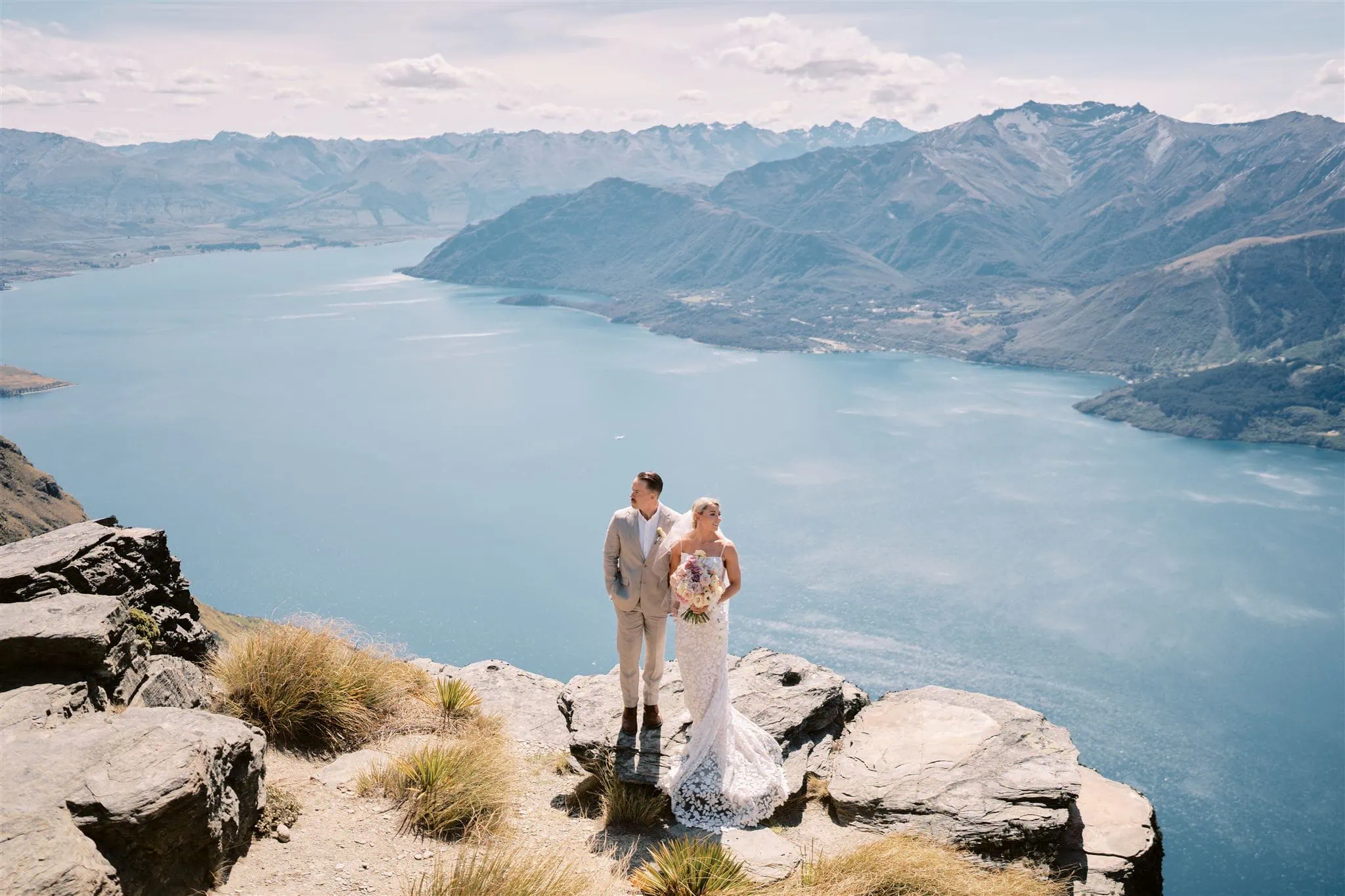 クイーンズタウン　テカポ　ニュージーランド　エロープメント 前撮り　フォトウェディング　結婚式 | ニュージーランドのクイーンズタウンにある山の崖の上で、美しい湖と山脈を見渡しながら手をつなぐ新郎新婦。