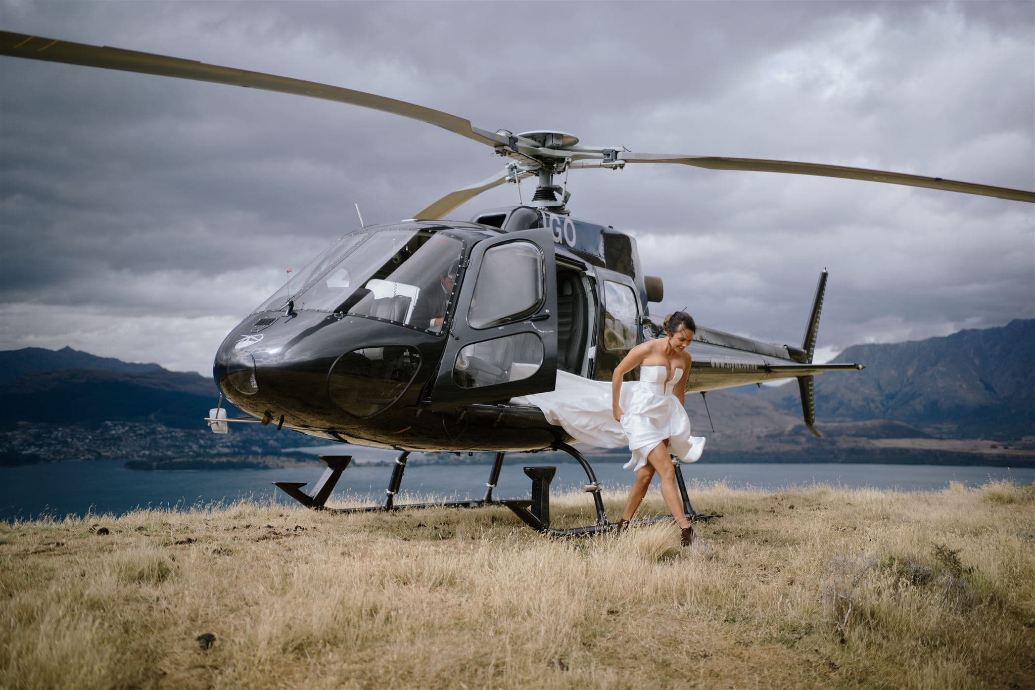 クイーンズタウン　テカポ　ニュージーランド　エロープメント 前撮り　フォトウェディング　結婚式 | 嵐の空と湖を背景に、白いドレスを着た女性がクイーンズタウンの草に覆われた丘の上でヘリコプターから走って逃げている。
