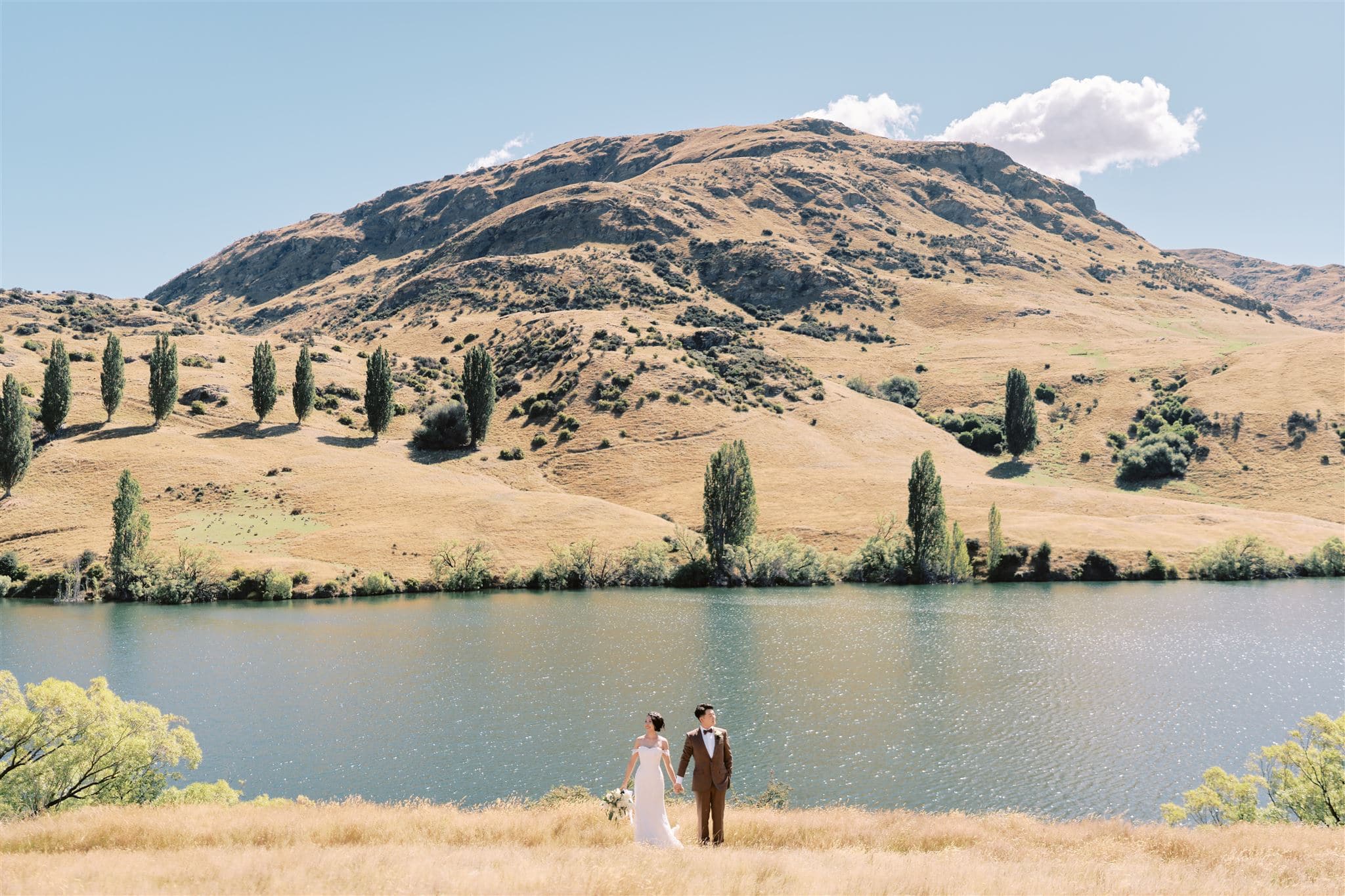 クイーンズタウン　テカポ　ニュージーランド　エロープメント 前撮り　フォトウェディング　結婚式 | クイーンズタウンの静かな湖のそばにカップルが立っています。背景には、澄み切った青空の下、木々が点在する大きな草の丘があります。