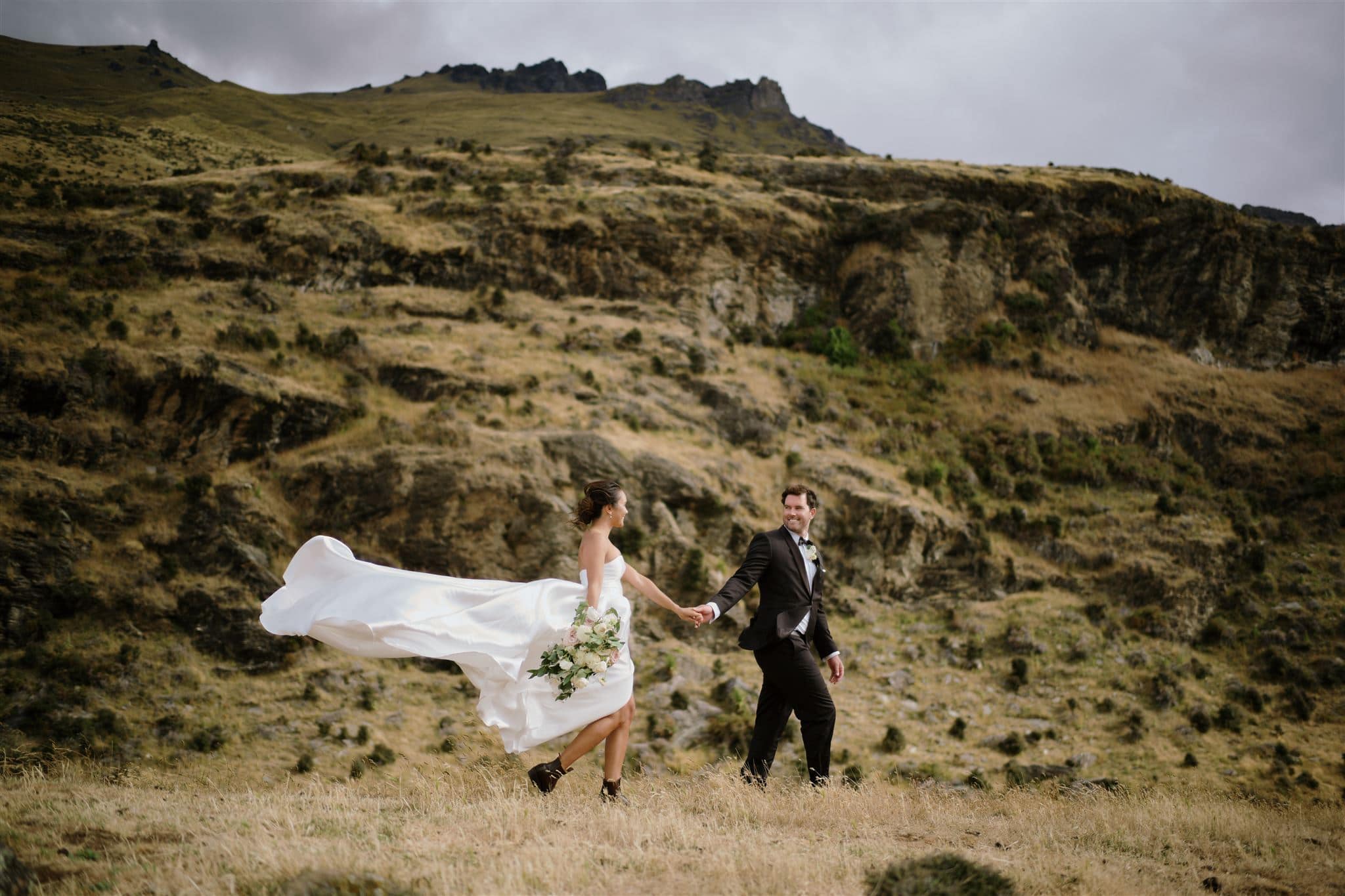 クイーンズタウン　テカポ　ニュージーランド　エロープメント 前撮り　フォトウェディング　結婚式 | ニュージーランドのクイーンズタウンで、花嫁と花婿が手をつなぎ、花嫁のドレスが風になびき、岩だらけの丘を背景にした険しい風景の中を楽しそうに歩いています。