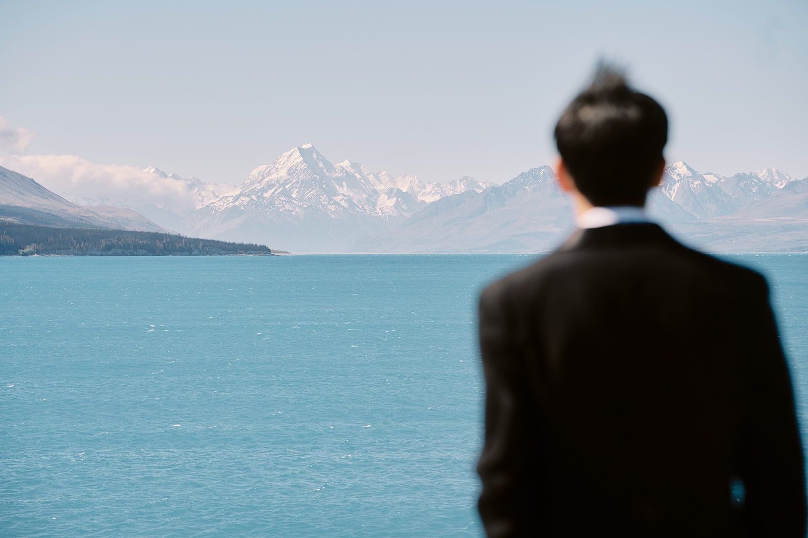 クイーンズタウン　テカポ　ニュージーランド　エロープメント 前撮り　フォトウェディング　結婚式 | ニュージーランドのテカポ湖の大きな青い湖の向こうに遠くの山を眺めるウェディングスーツを着た男性。