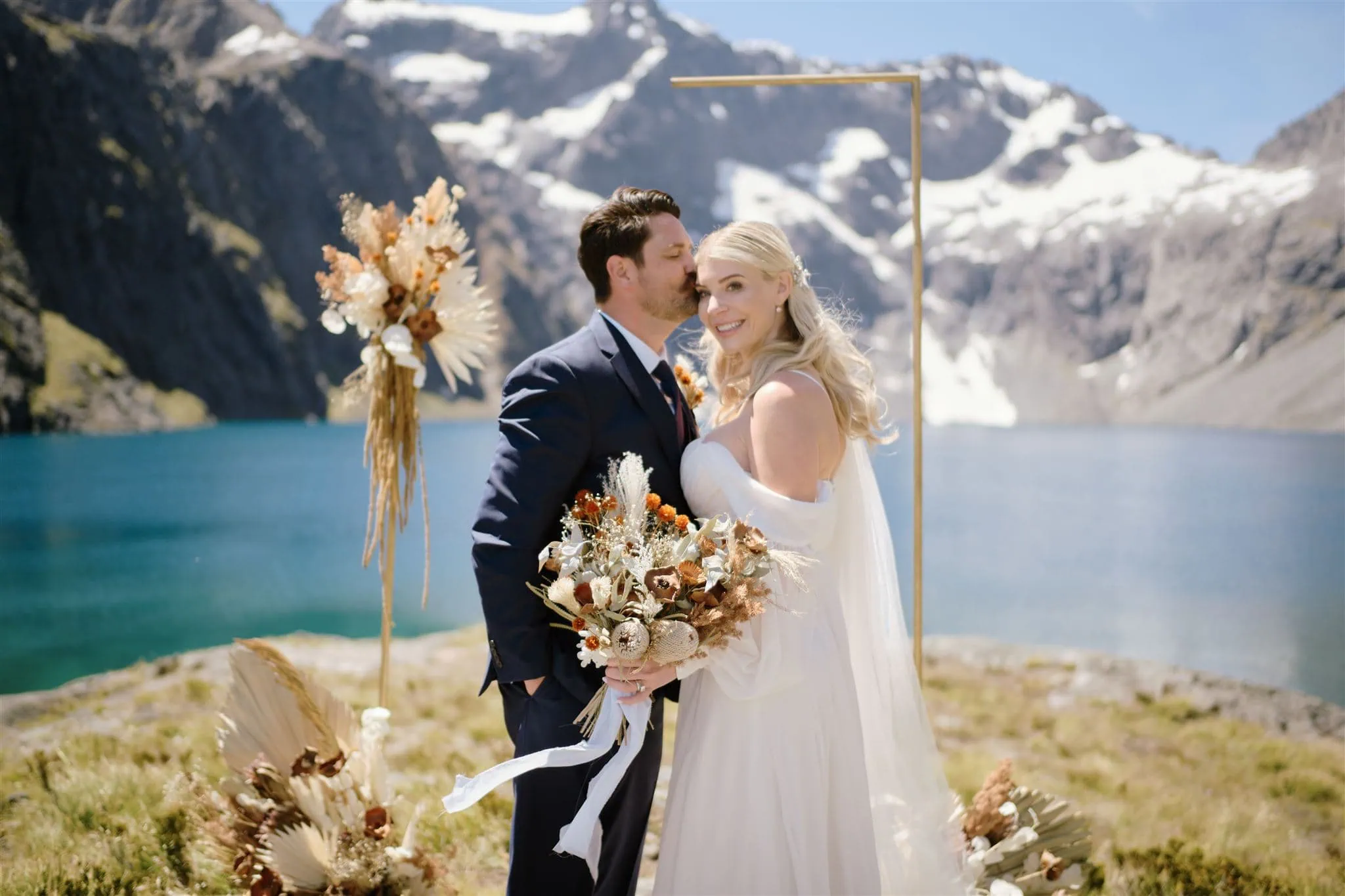 クイーンズタウン　テカポ　ニュージーランド　エロープメント 前撮り　フォトウェディング　結婚式 | クイーンズタウンの湖畔でカメラに向かって微笑む新婚カップル。花嫁はブーケを持ち、新郎はネイビーのスーツを着ています。
