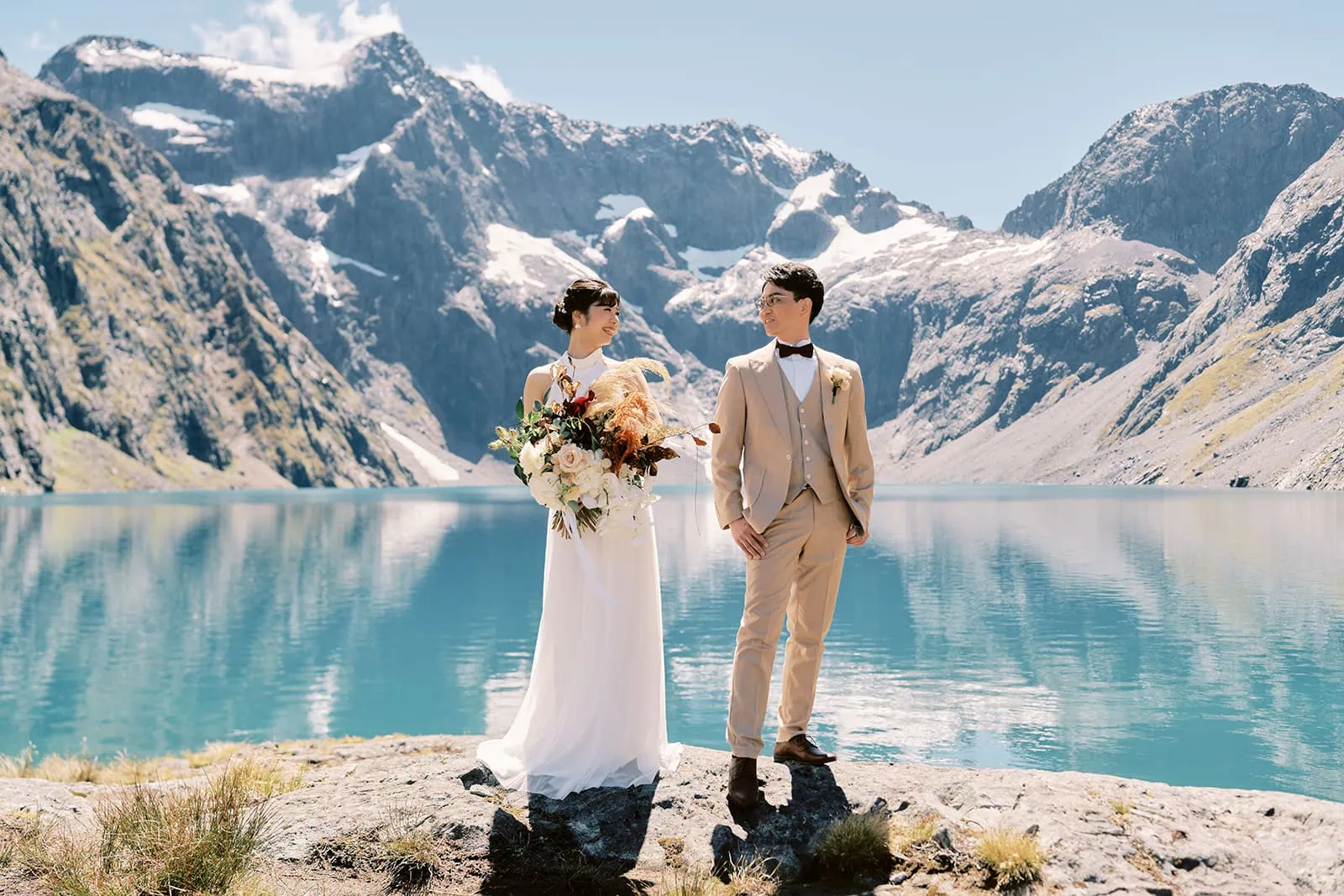 クイーンズタウン　テカポ　ニュージーランド　エロープメント 前撮り　フォトウェディング　結婚式 | ウェディングドレスを着たカップルが、雪をかぶった山頂を背景にした山の湖のそばに立って、クイーンズタウンウェディングを祝っています。